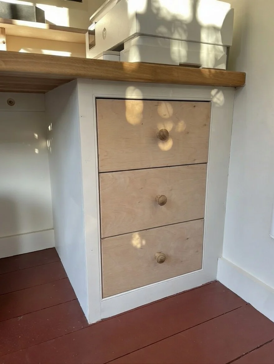 A white wooden kitchen cabinet with three drawers, each with a round wooden knob, against a wall with a wooden countertop. Part of a white appliance is visible on the countertop, and the floor is redwood.