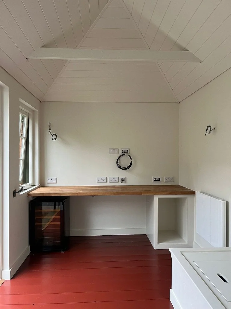 Empty room with a wooden countertop, a small wine refrigerator, electrical outlets, and exposed wiring on the walls, with a sloped ceiling and a window on the left side.