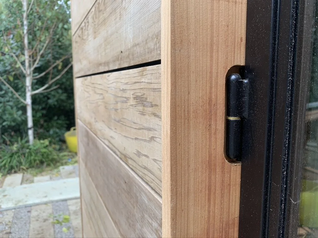 Close-up of a black hinge on a wooden wall next to a glass door, with a garden in the background.