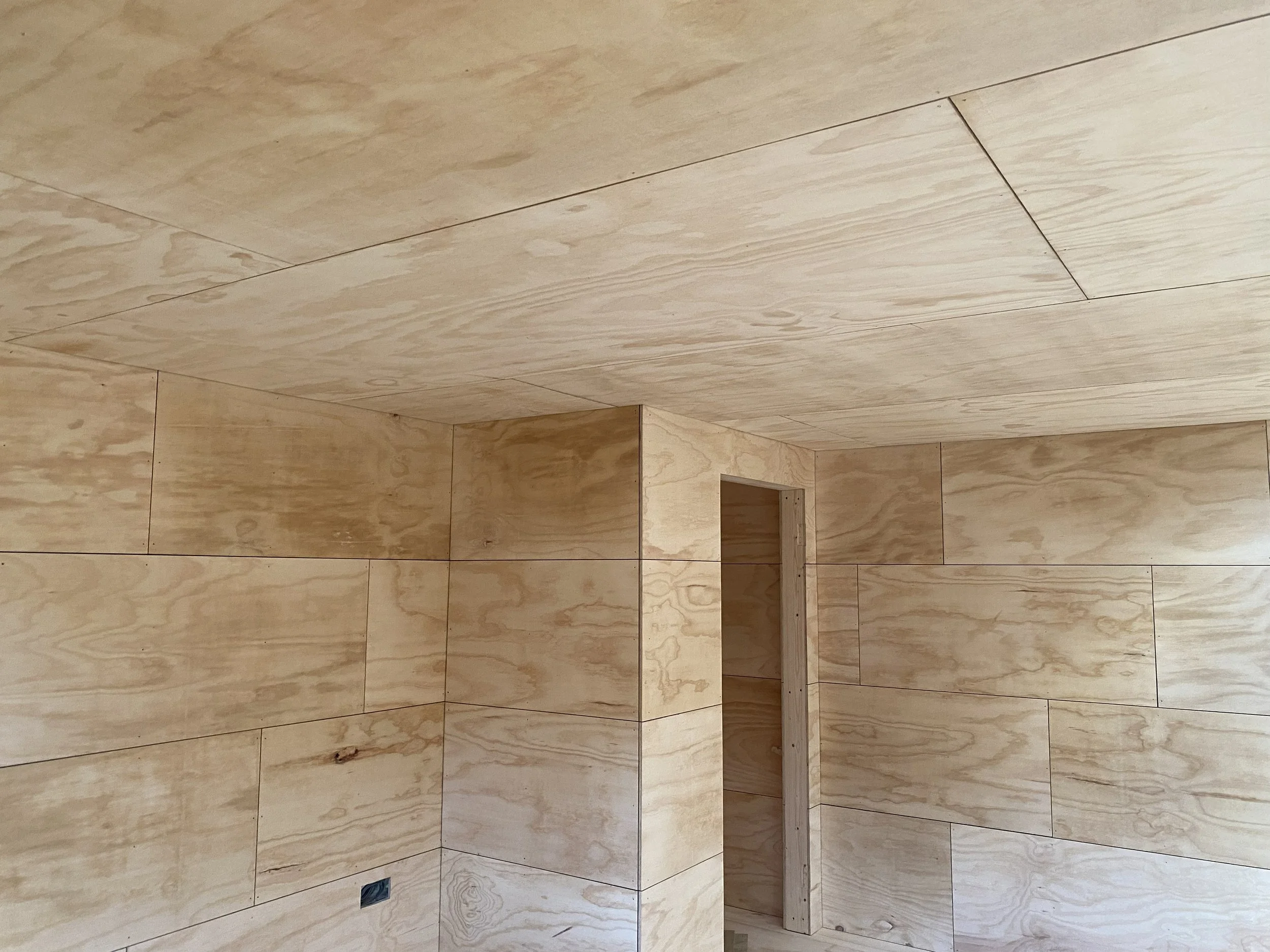 Interior of a room with unfinished wooden walls and ceiling, including a small doorway opening, and a power outlet on the lower wall.