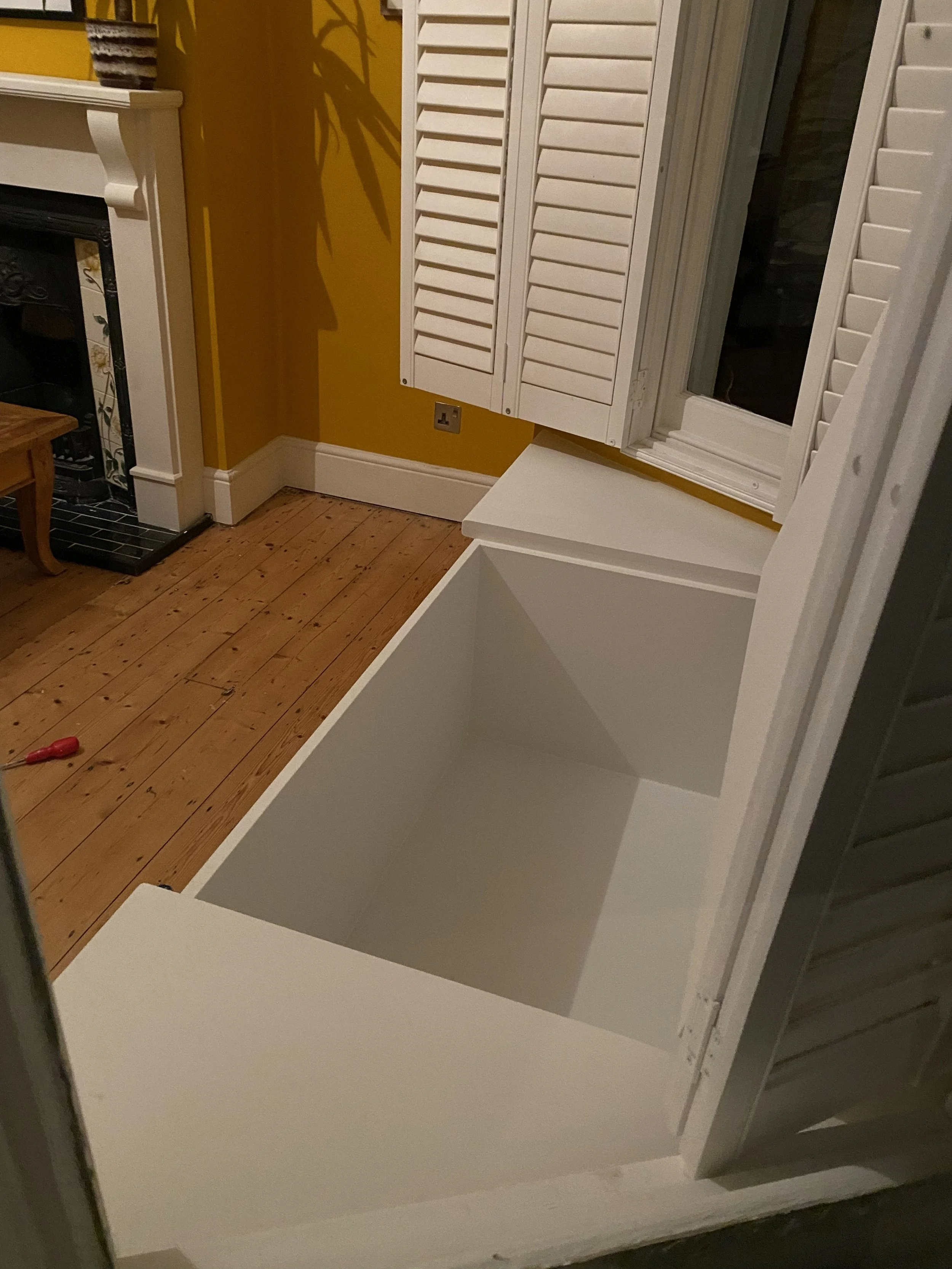 The image shows a white storage box or cabinet with a window and white shutters on top, set against a yellow wall with a wood floor.