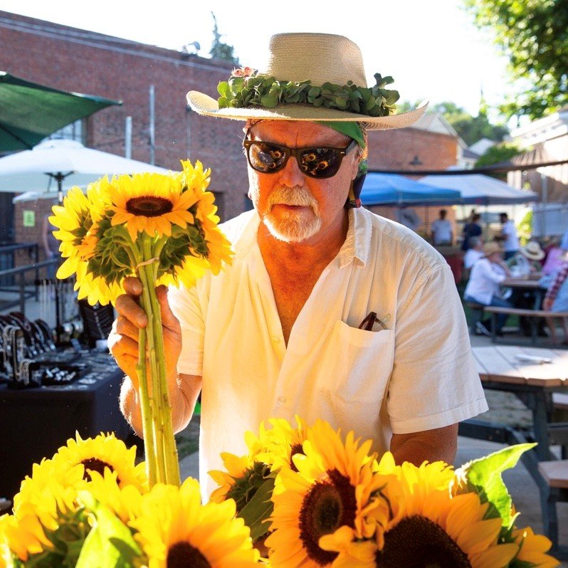Ron Antone smiling at the Plymouth farmers market, holding bright sunflowers and wearing a straw hat