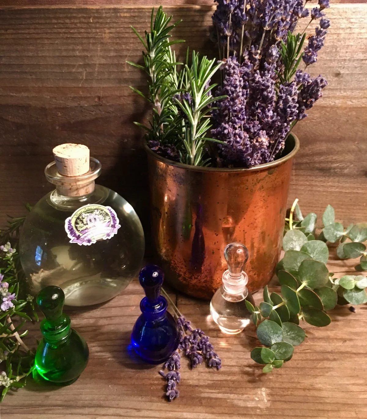 Lavender and rosemary in a copper mug beside small glass essential oil bottles on a warm wood surface