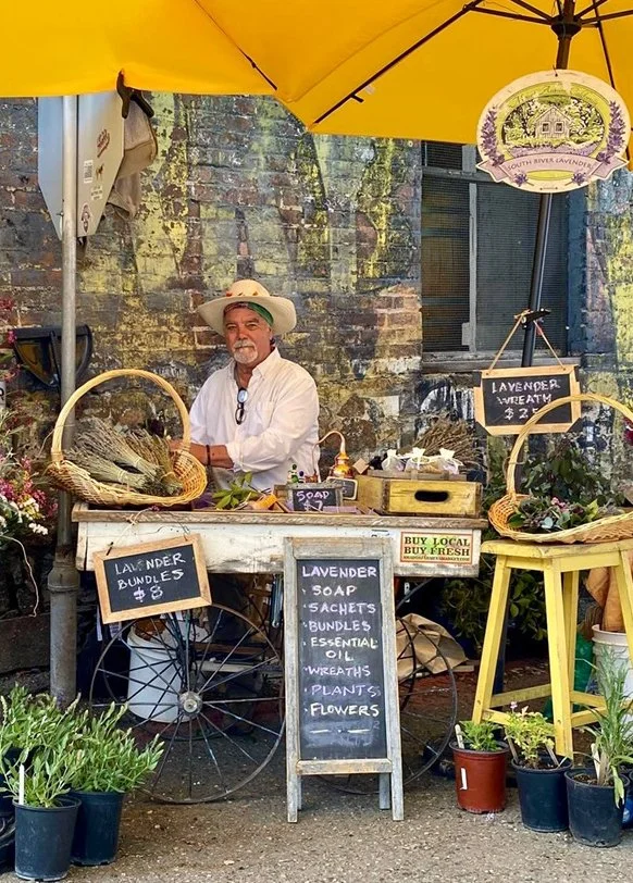 Ron Antone at his market stand under a yellow umbrella with lavender bundles, baskets, and a chalkboard menu
