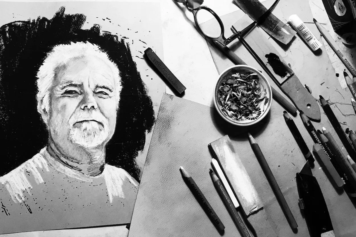 Ron Antone's charcoal self-portrait in progress on a drawing table surrounded by pencils and charcoal sticks