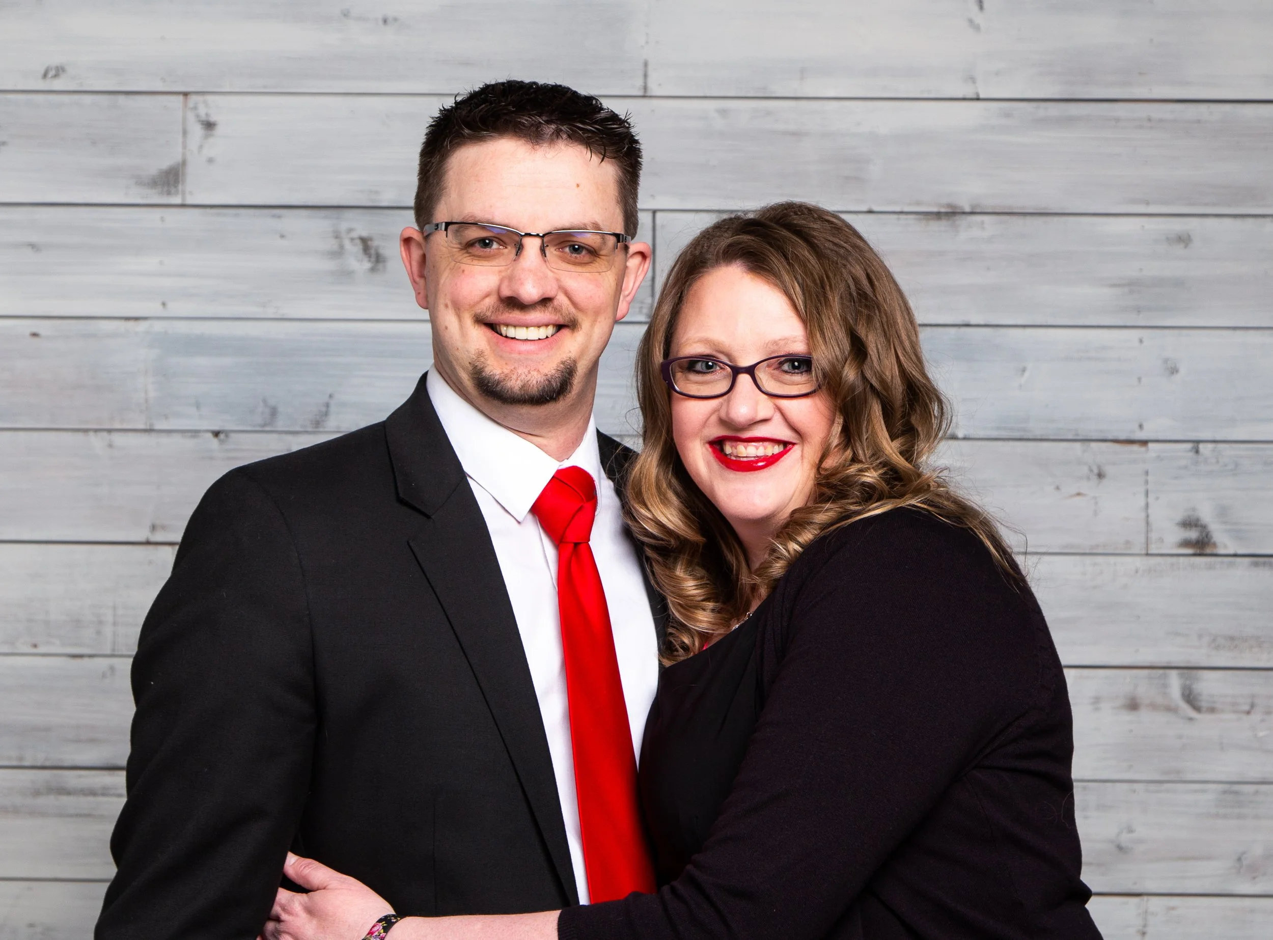 A smiling couple posing together in front of a light gray wooden wall. The man is wearing a black suit, white shirt, and red tie with glasses, and the woman is wearing a black top and glasses, with curly hair and bright red lipstick.