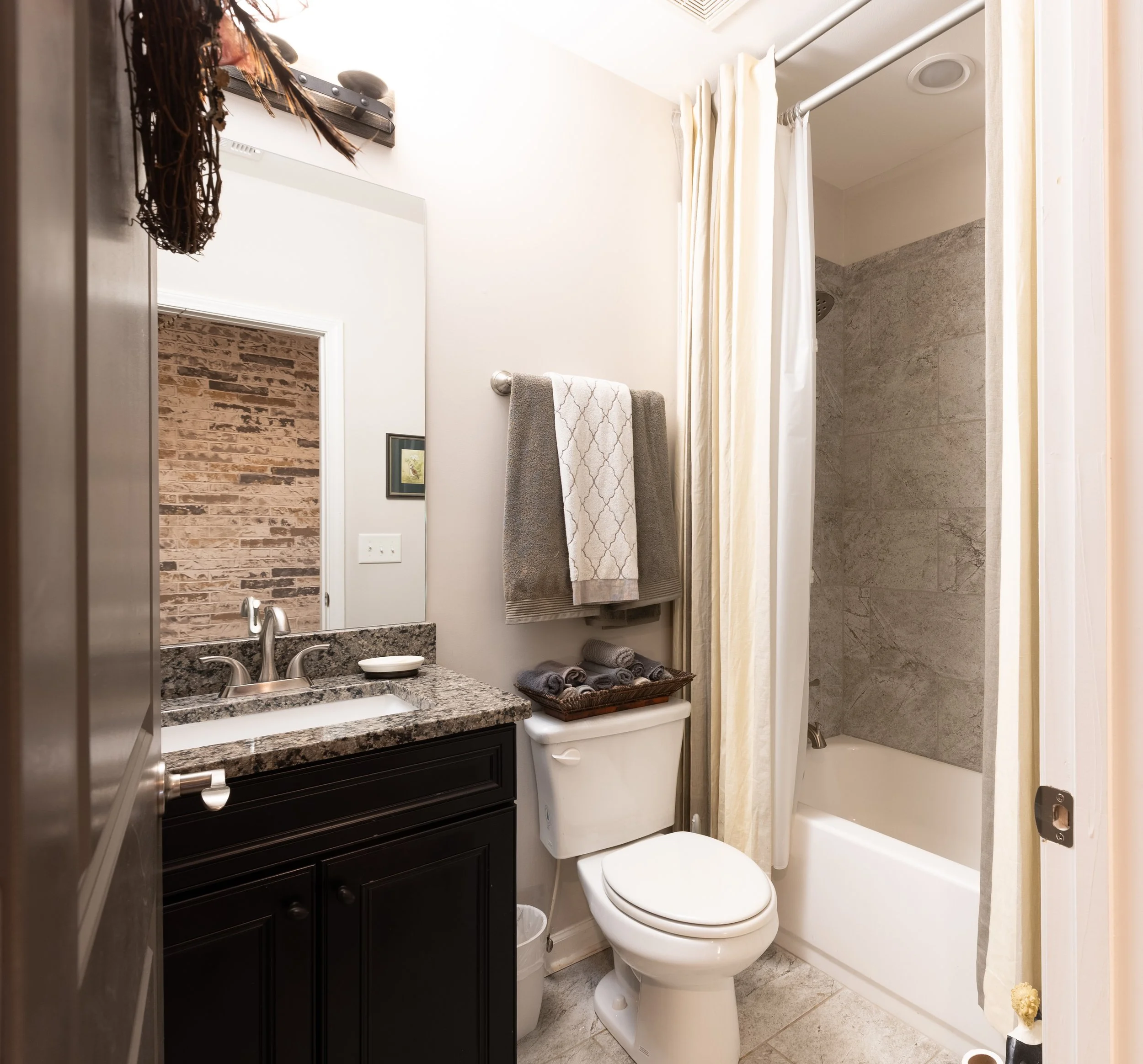 Small bathroom with a granite countertop sink, a mirror, a towel rack with two towels, a shelf with rolled towels above the toilet, a shower with a curtain, and a small trash bin on the floor.