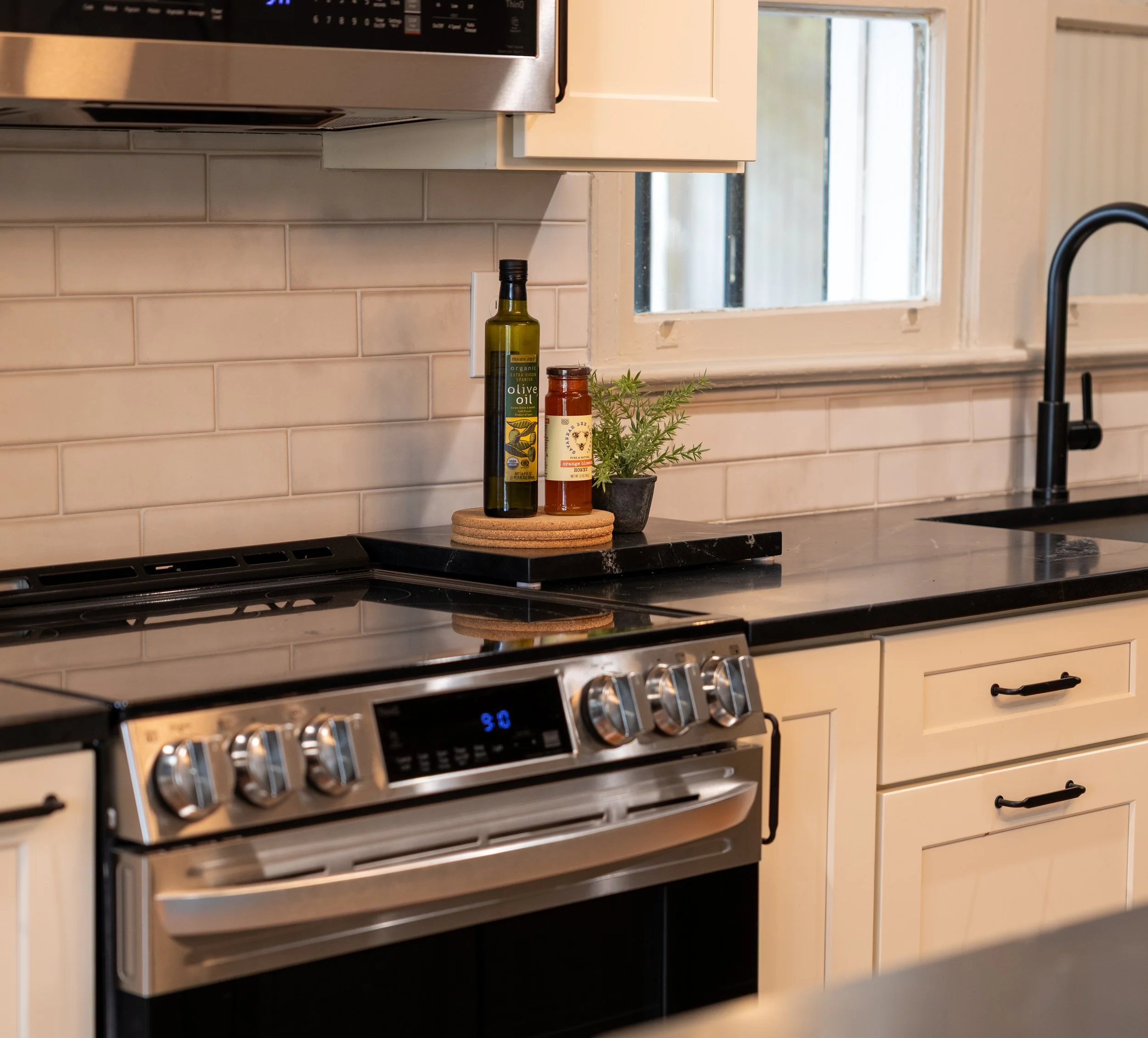 Kitchen countertop with olive oil and hot sauce bottles, a small potted plant, black countertop, stove, and window with white frame in the background.