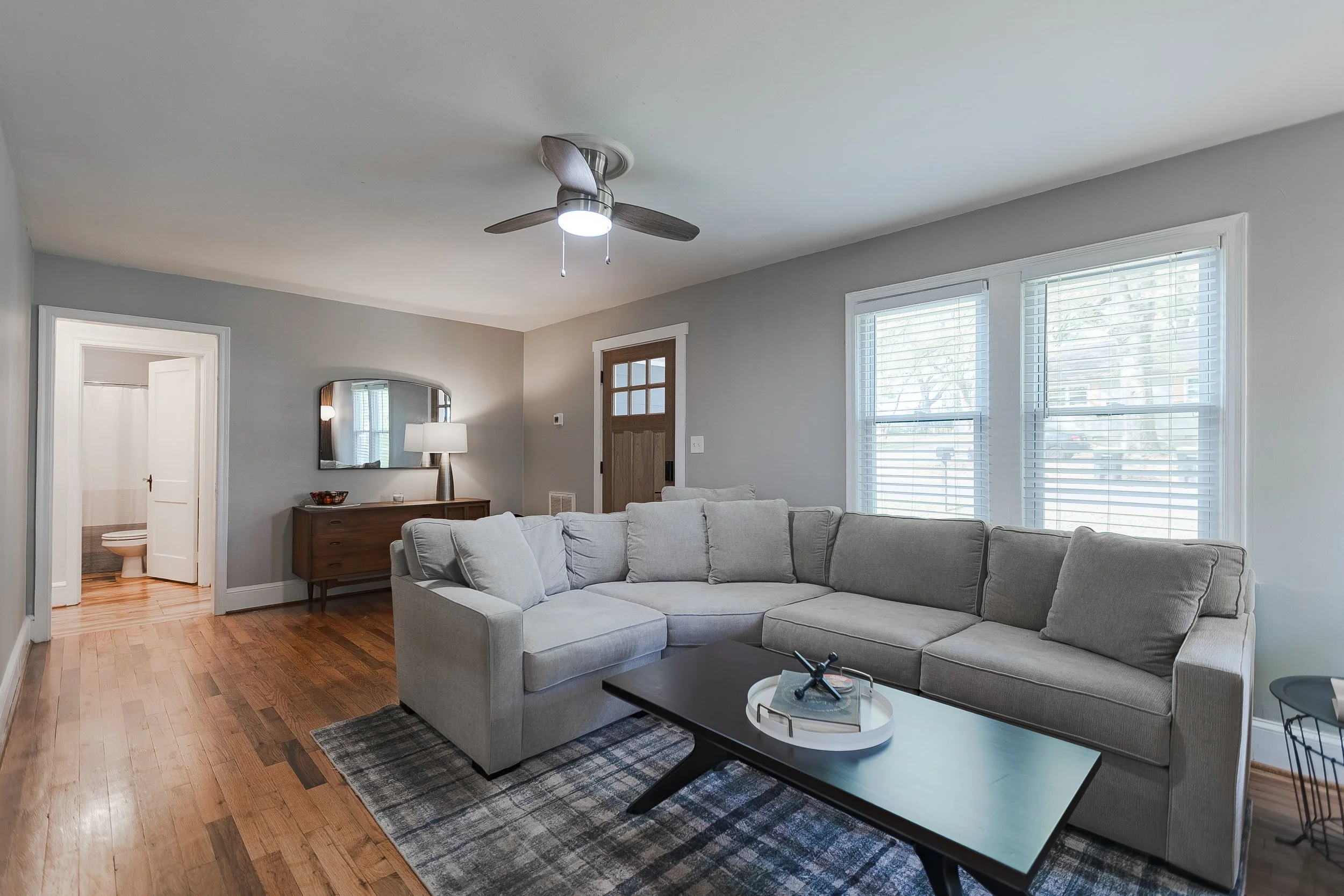 Living room with a beige sectional sofa, a black coffee table, a gray area rug, a wooden sideboard with a mirror and a lamp, four windows with white blinds, a ceiling fan, and a doorway leading to a bathroom.