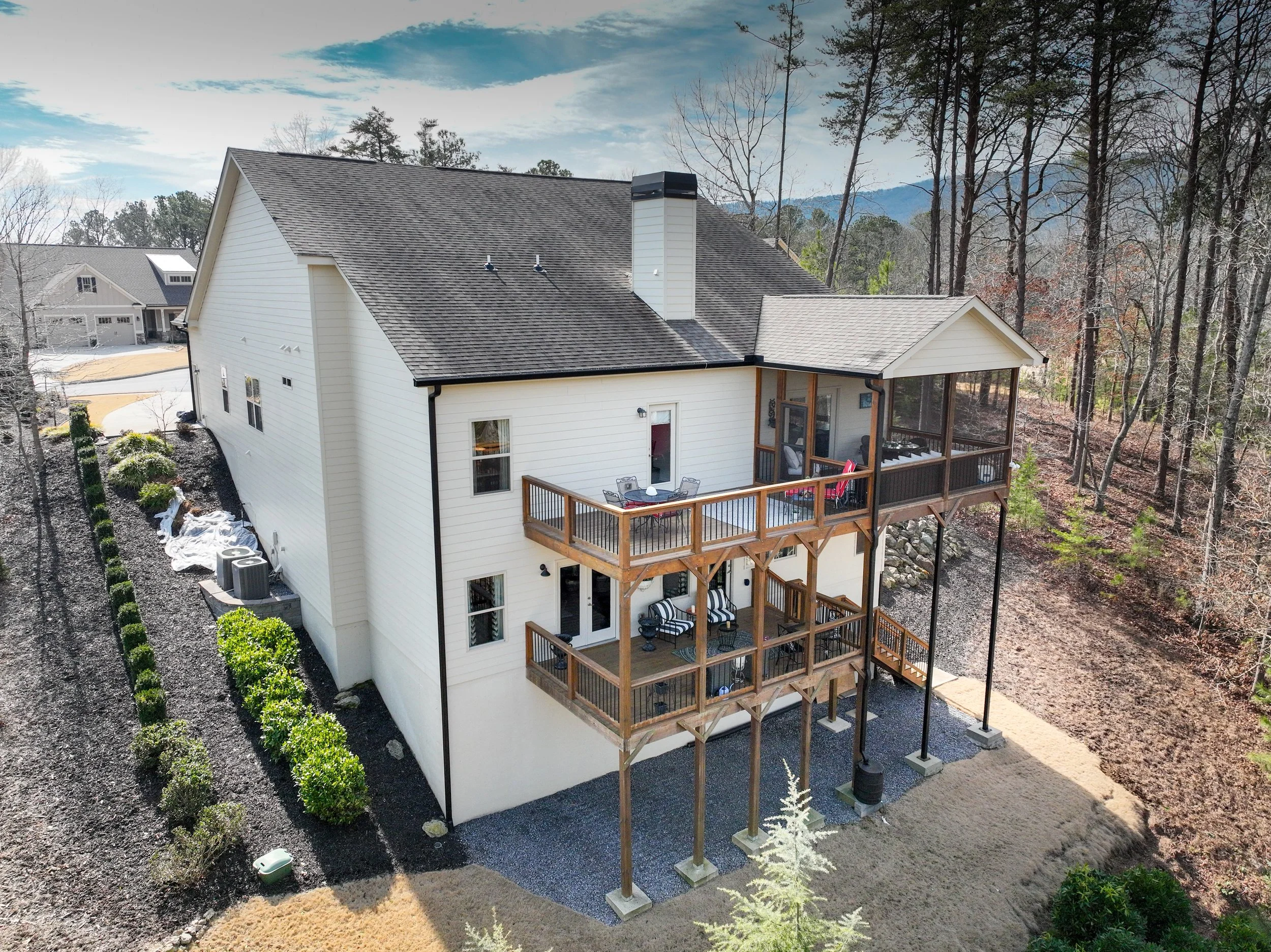 A multi-story house with two wooden decks, one on each level, in a rural wooded area with many leafless trees and a mountain in the background. The house has white siding, a shingled roof, and a screened porch. There is gravel on the ground beneath the decks, and landscaped bushes along the side of the house.