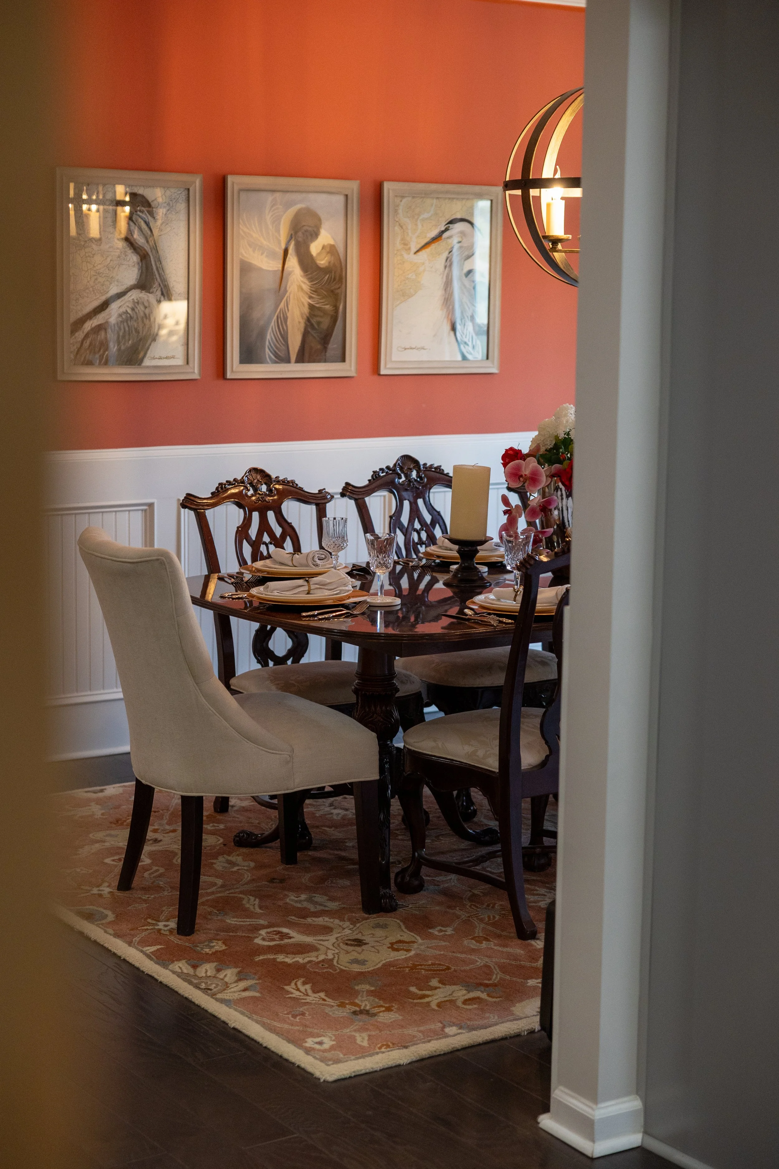 A dining room with a wooden table set for a meal, surrounded by ornate wooden chairs and beige upholstery. The table has a centerpiece with pink and white flowers and a large white candle. The room has a coral-colored wall with three framed bird illustrations and a modern spherical wall sconce lighting fixture.