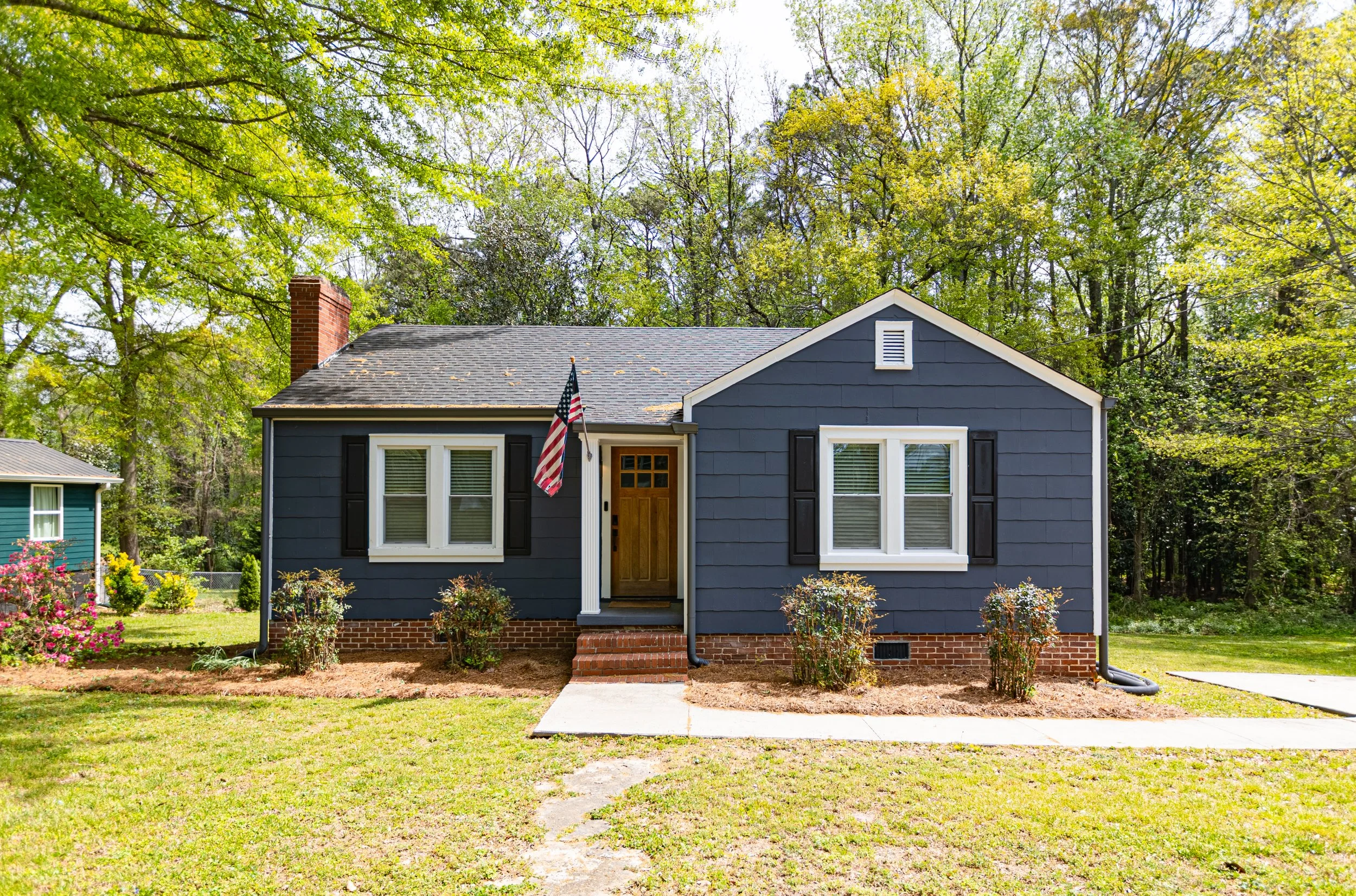 Front view of a dark blue house with white trim, black shutters, a brick foundation, and a small staircase leading to the front door, with a lawn and trees in the background.