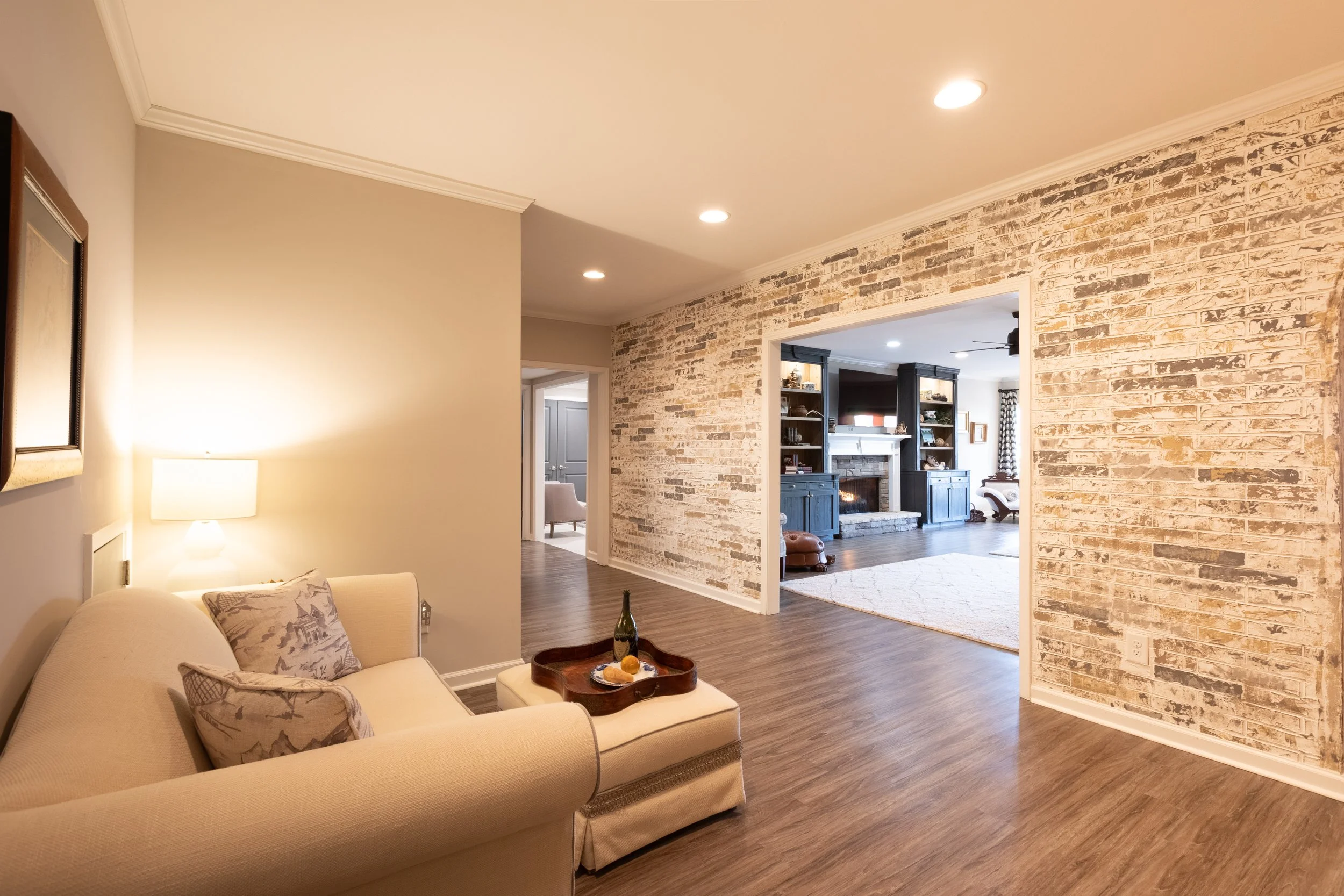 Living room with a beige sofa, pillows, a lamp, and a tray with a bottle and snacks, leading to a second room with dark wood cabinetry and a fireplace.