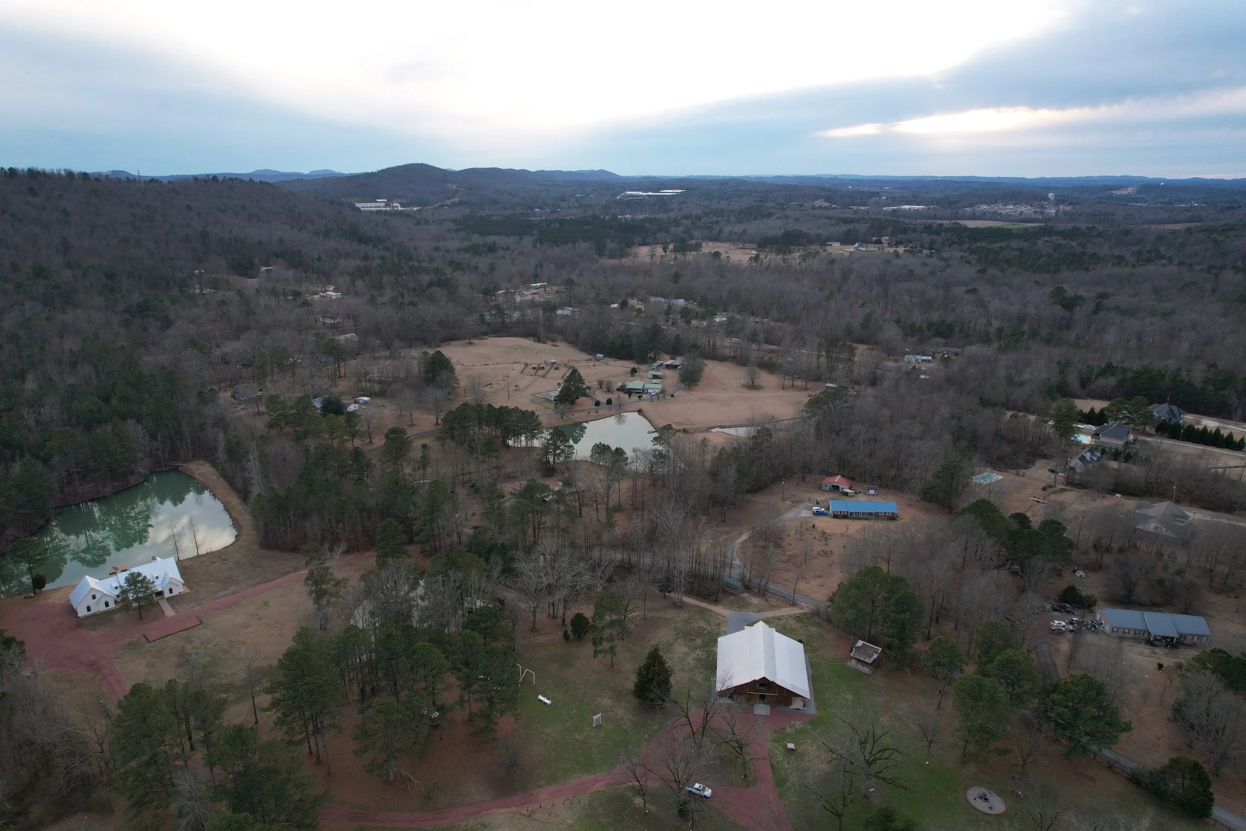 Aerial view of a rural landscape with lakes, trees, buildings, and hills in the distance on a cloudy day.