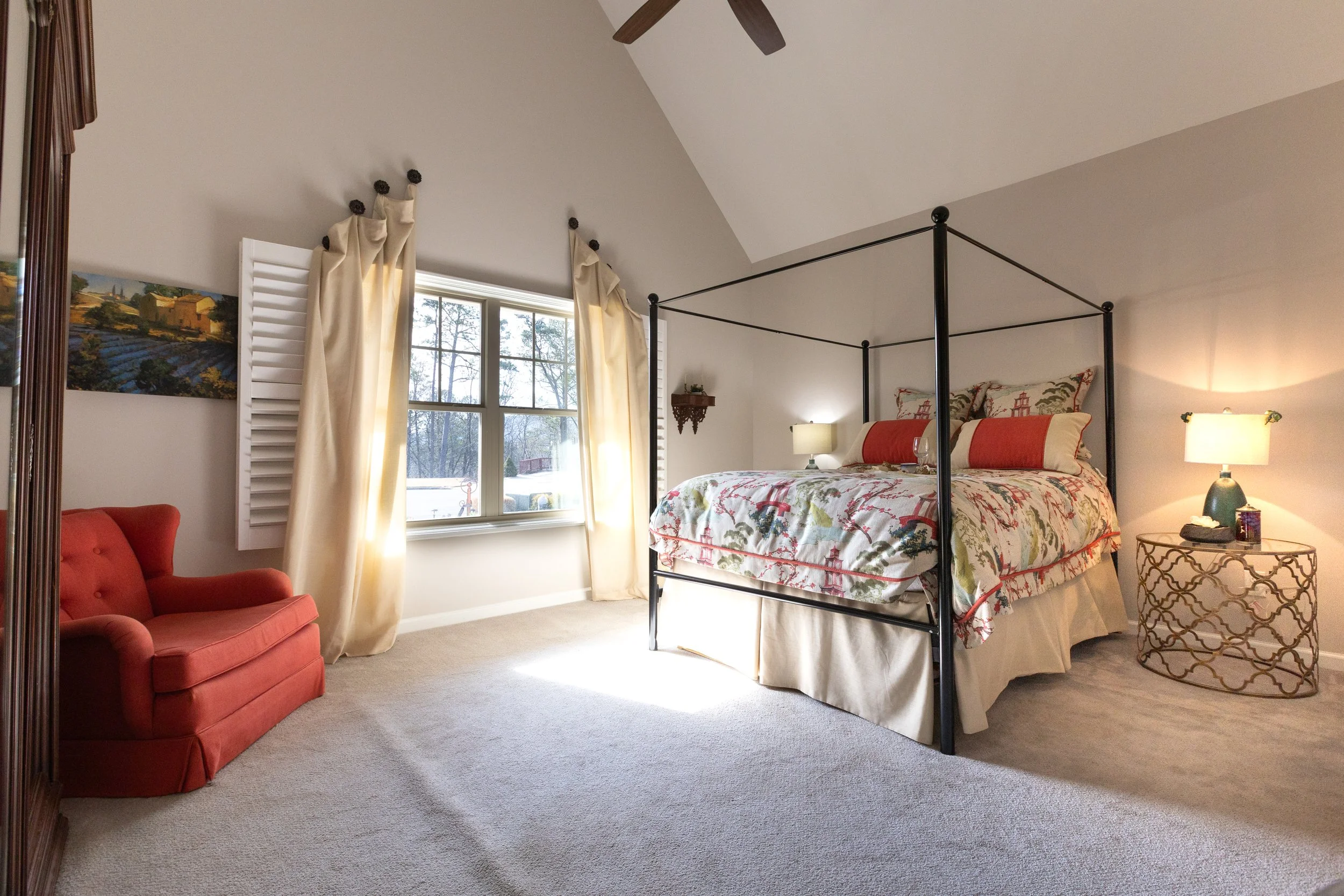 A cozy bedroom with a four-poster bed, patterned bedding, two nightstands with lamps, a red armchair, a large window with cream-colored curtains, beige carpet, and a painting on the wall.