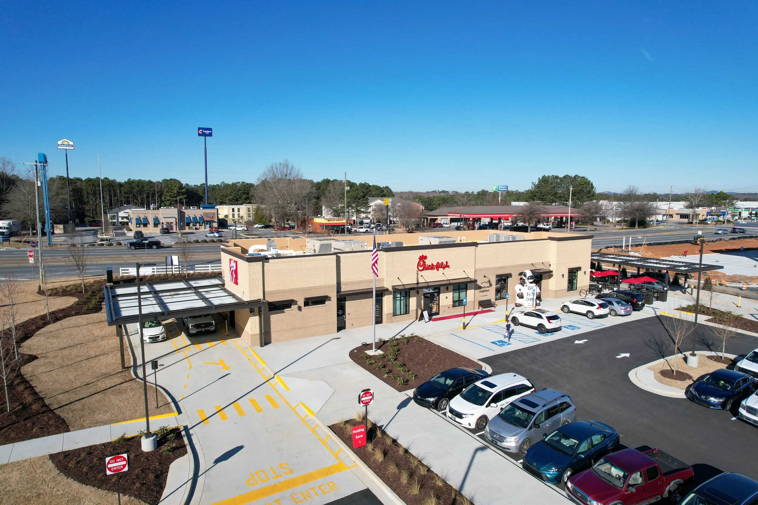 A Chick-fil-A restaurant with parking lot, drive-thru, and surrounding commercial area under a clear blue sky.