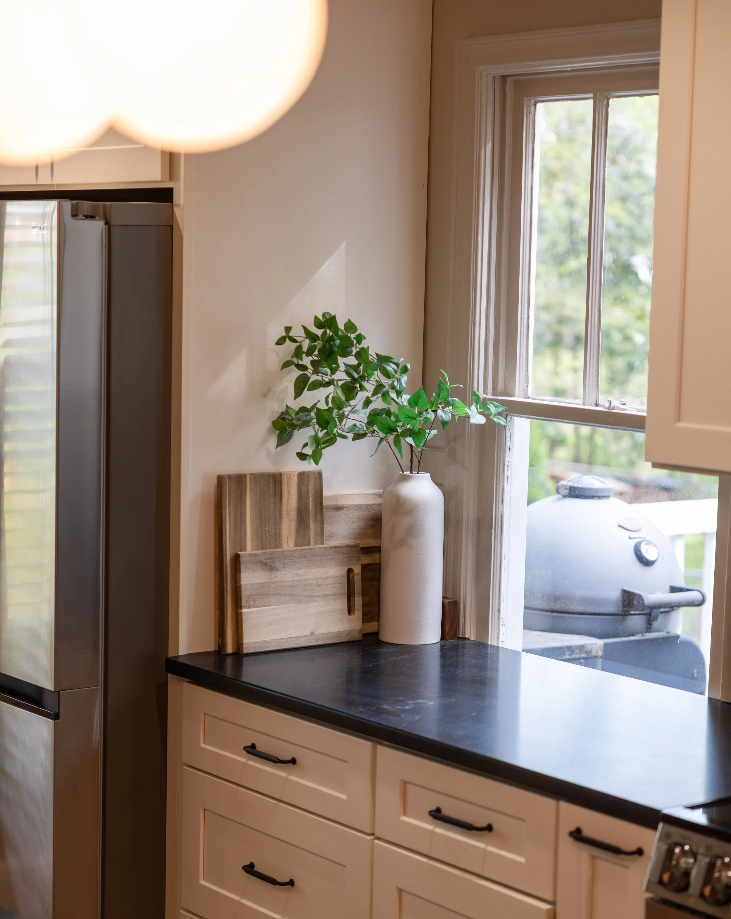 Kitchen countertop with a white vase holding green leaves, wooden cutting boards, and a window with a grill and a barbecue grill outside.