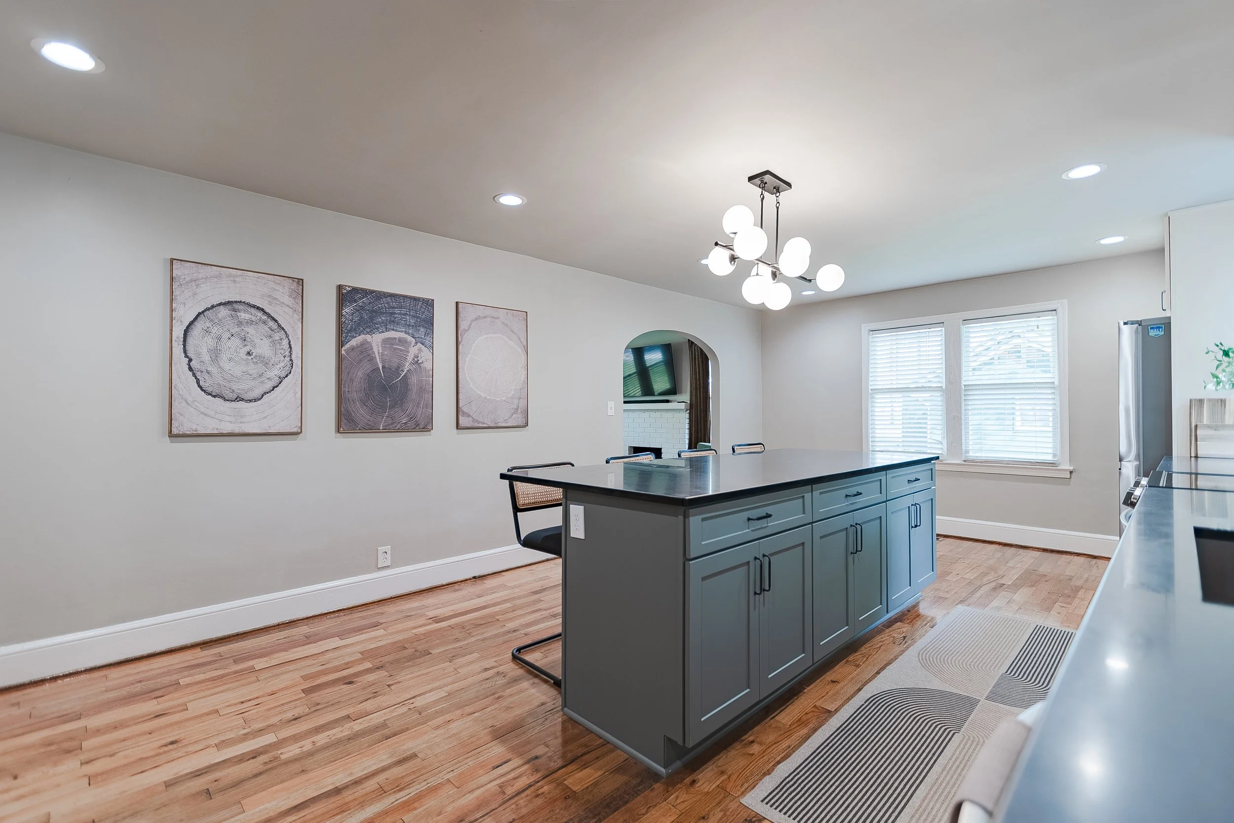 Modern kitchen with gray island, wooden floors, two windows with white blinds, wall art of tree cross-sections, and a geometric rug.