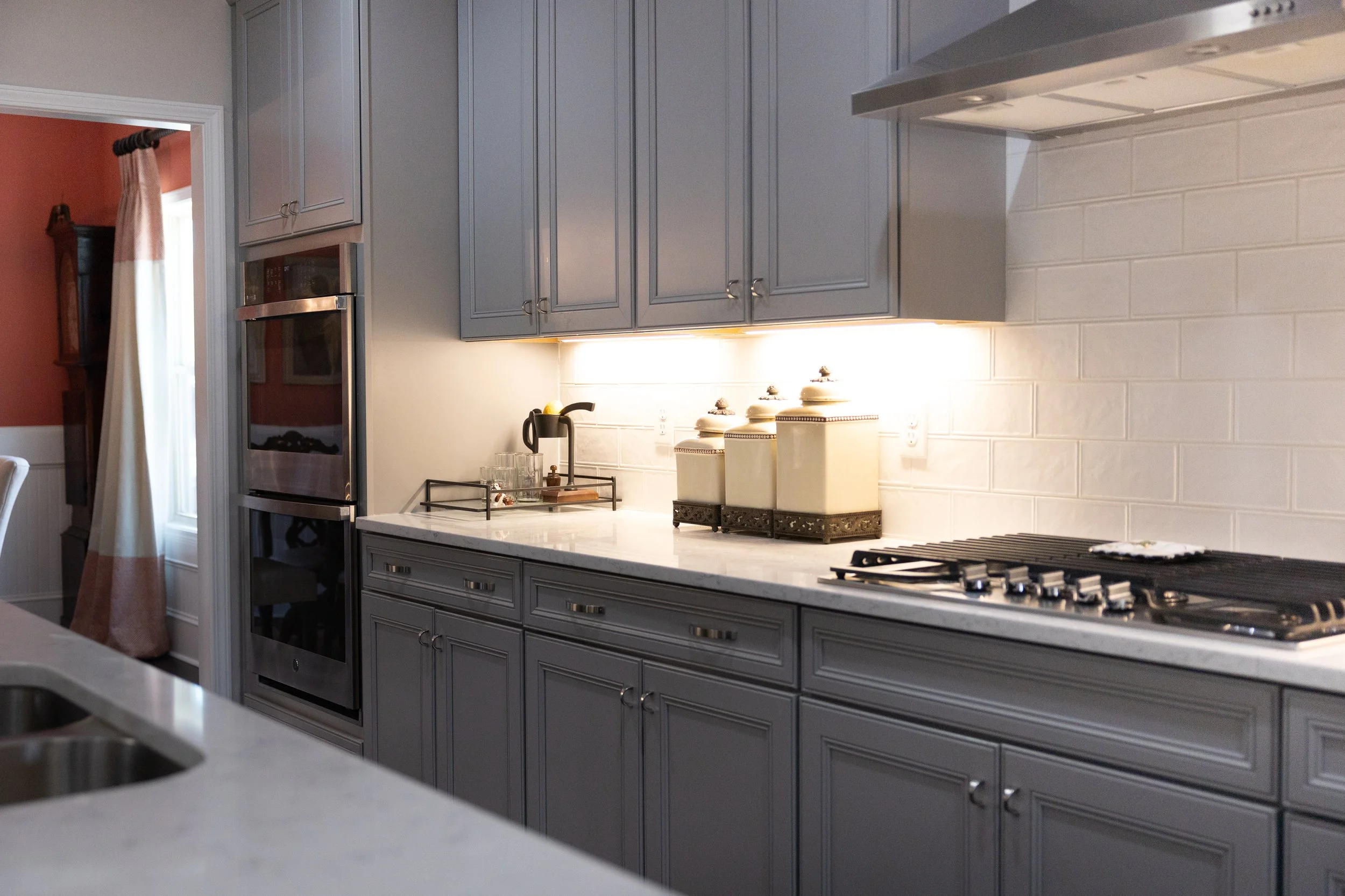 Kitchen with gray cabinets, white subway tile backsplash, gas stove, and built-in oven, with beige canisters on the countertop and a black tray holding glasses near the wall.