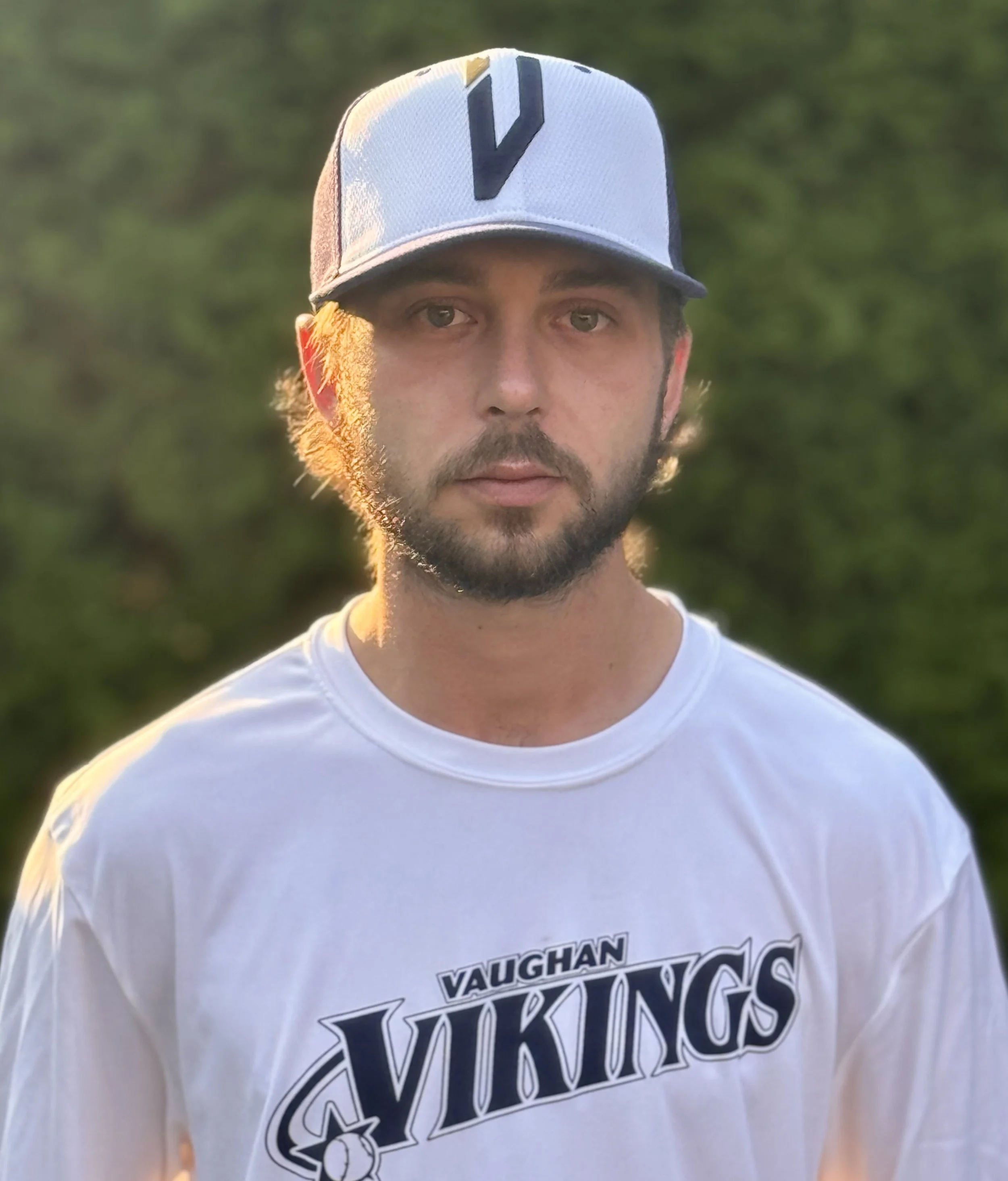 A young man with a beard and mustache wearing a white Vaughn Vikings sports jersey and a white and black baseball cap, standing outdoors with a blurred green background.