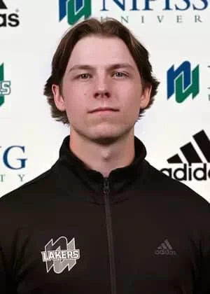A young man with medium-length brown hair wearing a black Adidas jacket with a LAKERS logo, standing in front of a backdrop with university and sponsor logos.
