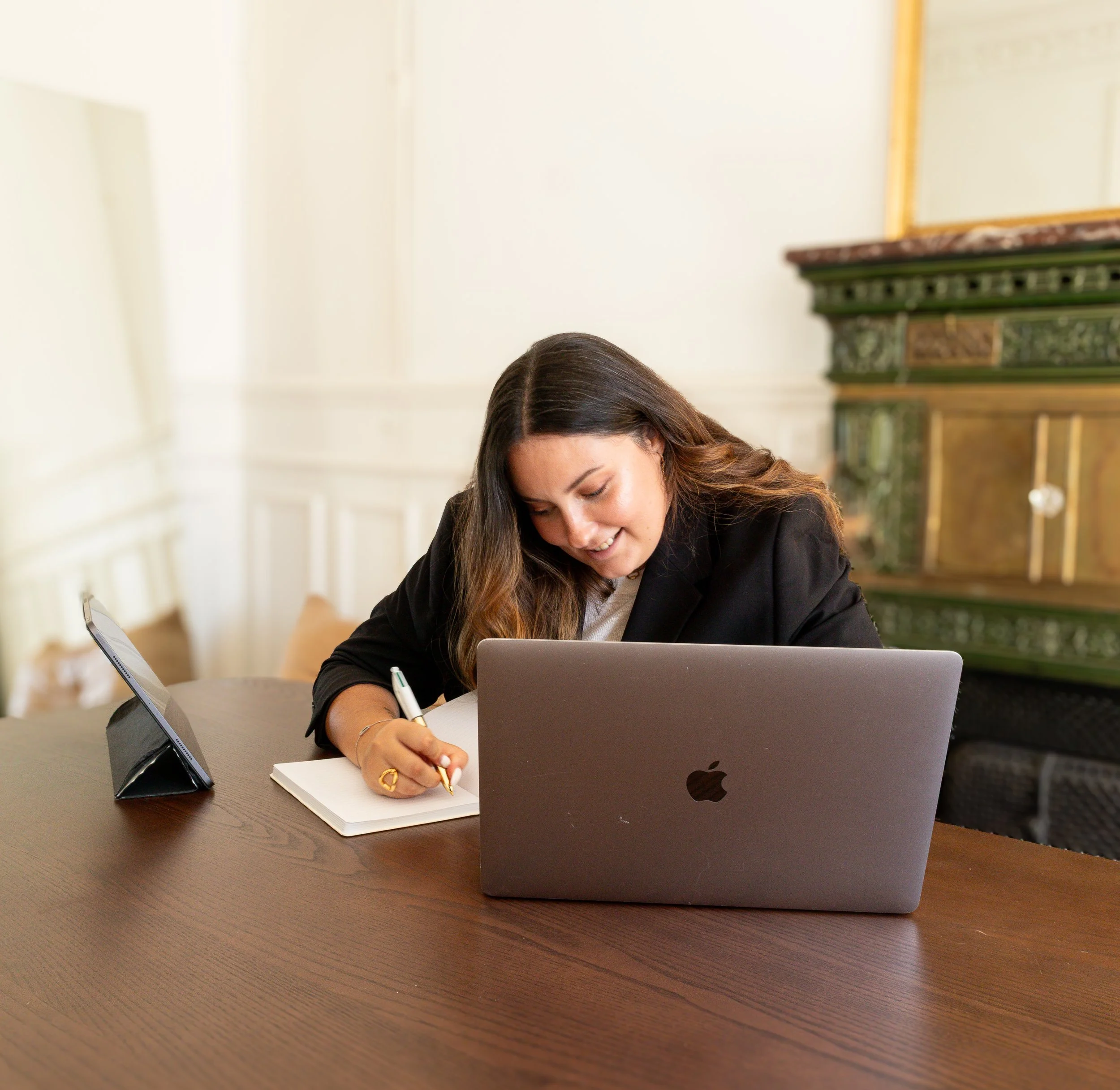 Une femme avec des cheveux bruns foncés, portant un costume noir, écrit dans un carnet blanc avec un stylo. Elle est assise à une table en bois avec un ordinateur portable gris clair devant elle. Sur la table, il y a un support de téléphone portable en forme de triangle noir. La pièce a des murs blancs avec des moulures et une cheminée en marbre avec des livres dessus.