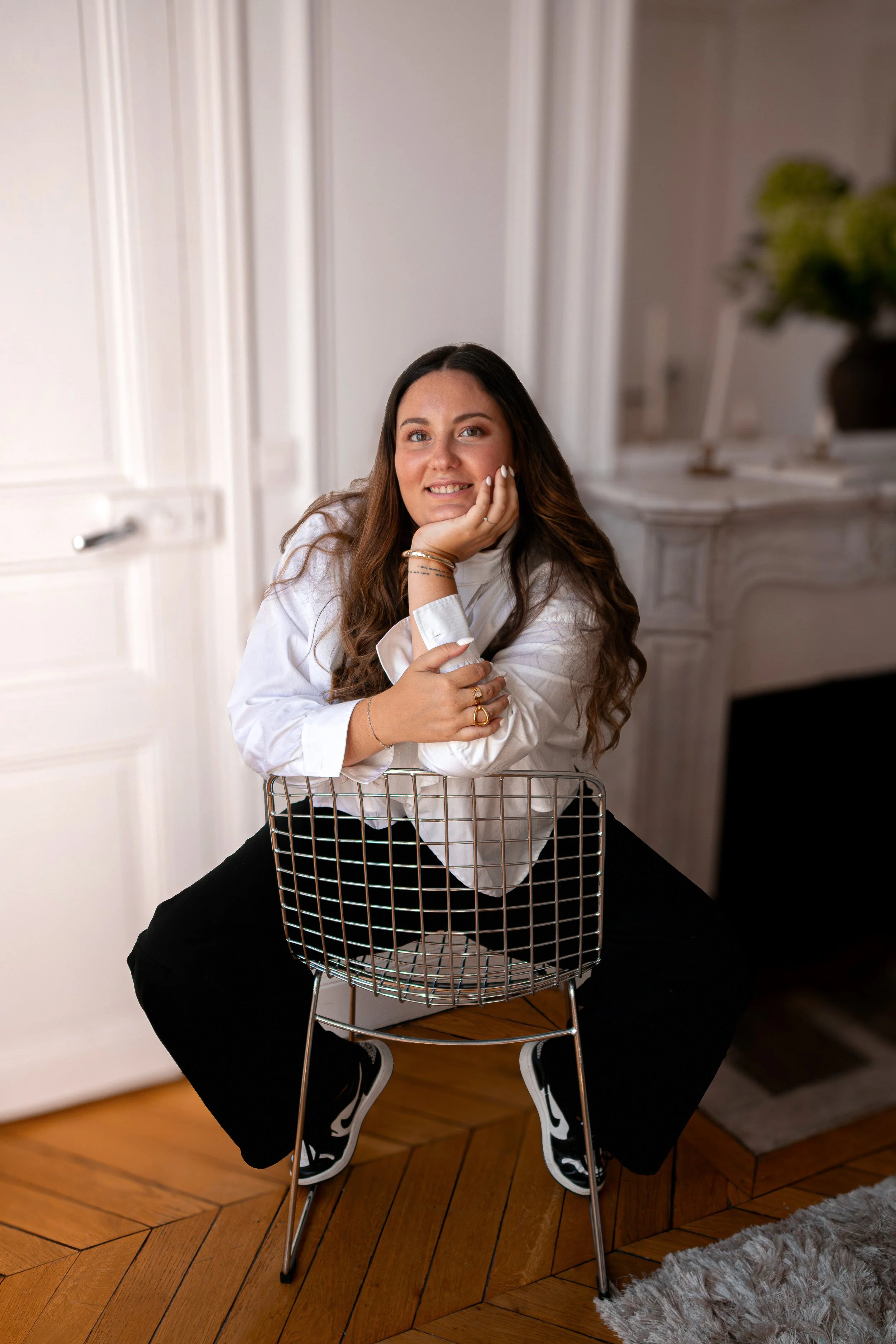 Une femme avec de longs cheveux bruns, assise sur une chaise en métal, posant avec le visage soutenu par sa main et souriant, dans un intérieur lumineux avec des murs clairs et une décoration moderne.
