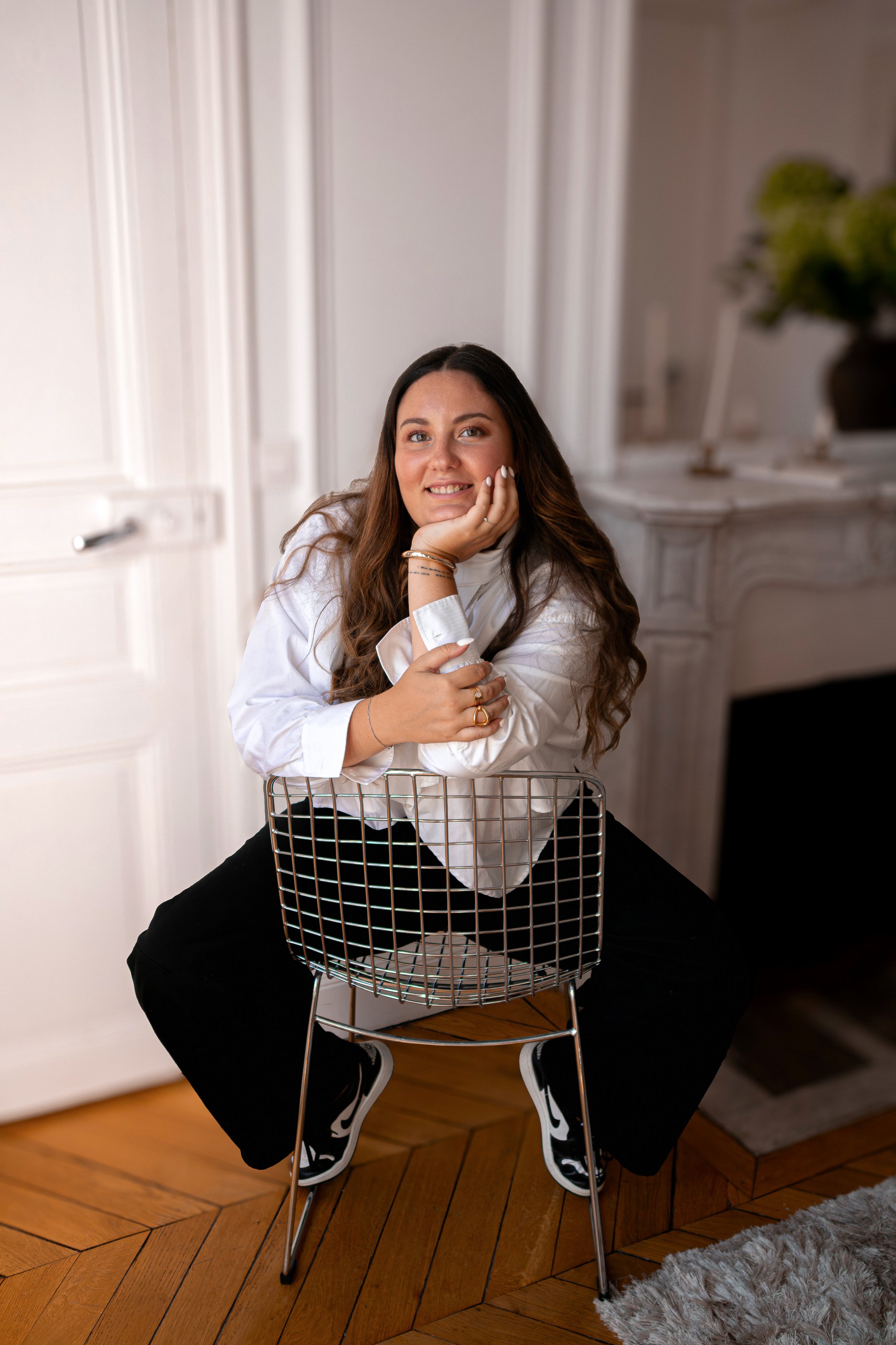 Une femme souriante avec des cheveux bruns longs, portant un chemisier blanc et un pantalon noir, repose sa tête sur sa main tout en étant assise sur une chaise en fil métallique dans une pièce lumineuse avec un parquet en bois et un tapis gris clair.