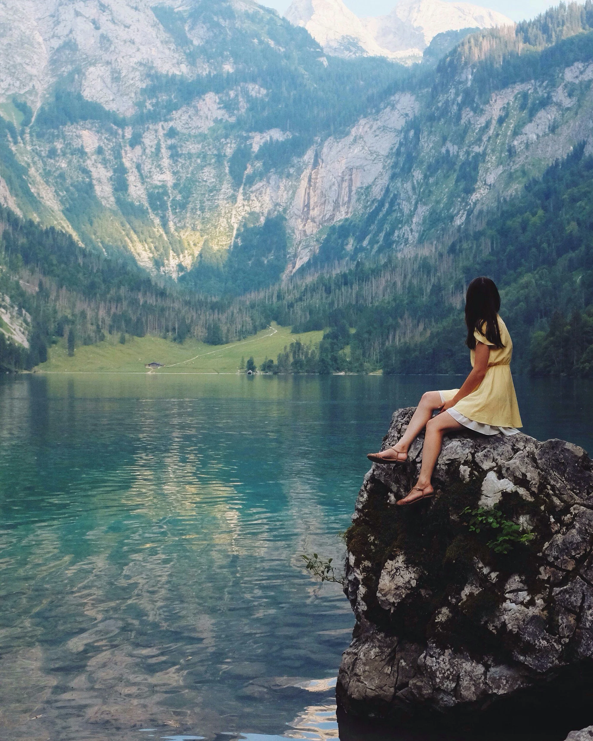 A girl in a yellow dress sitting on a rock by a lake with mountains and trees in the background.