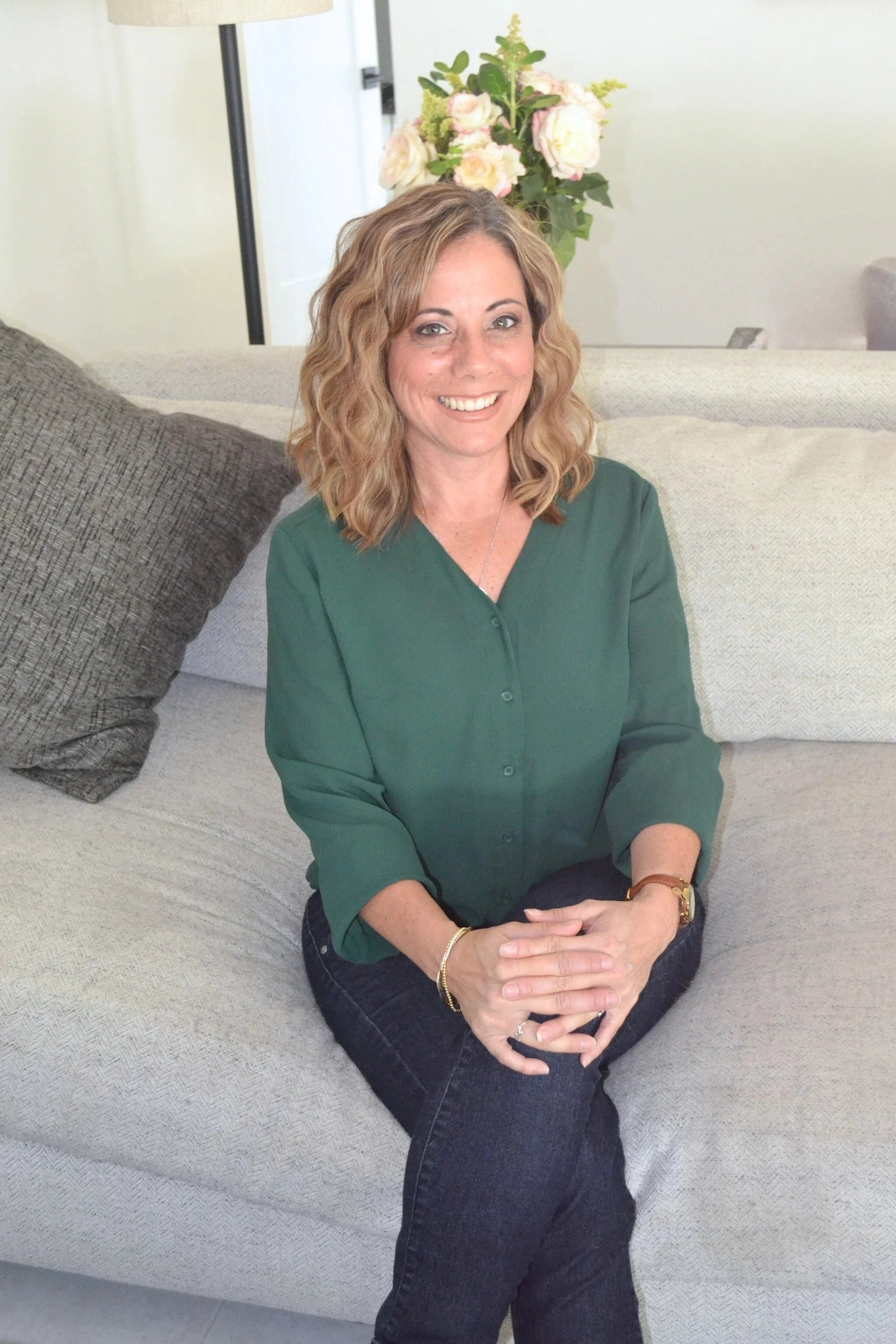A woman with wavy blonde hair wearing a green blouse and dark jeans, sitting on a beige sofa with her hands clasped, smiling at the camera. In the background, there is a bouquet of pink and white flowers on a table.
