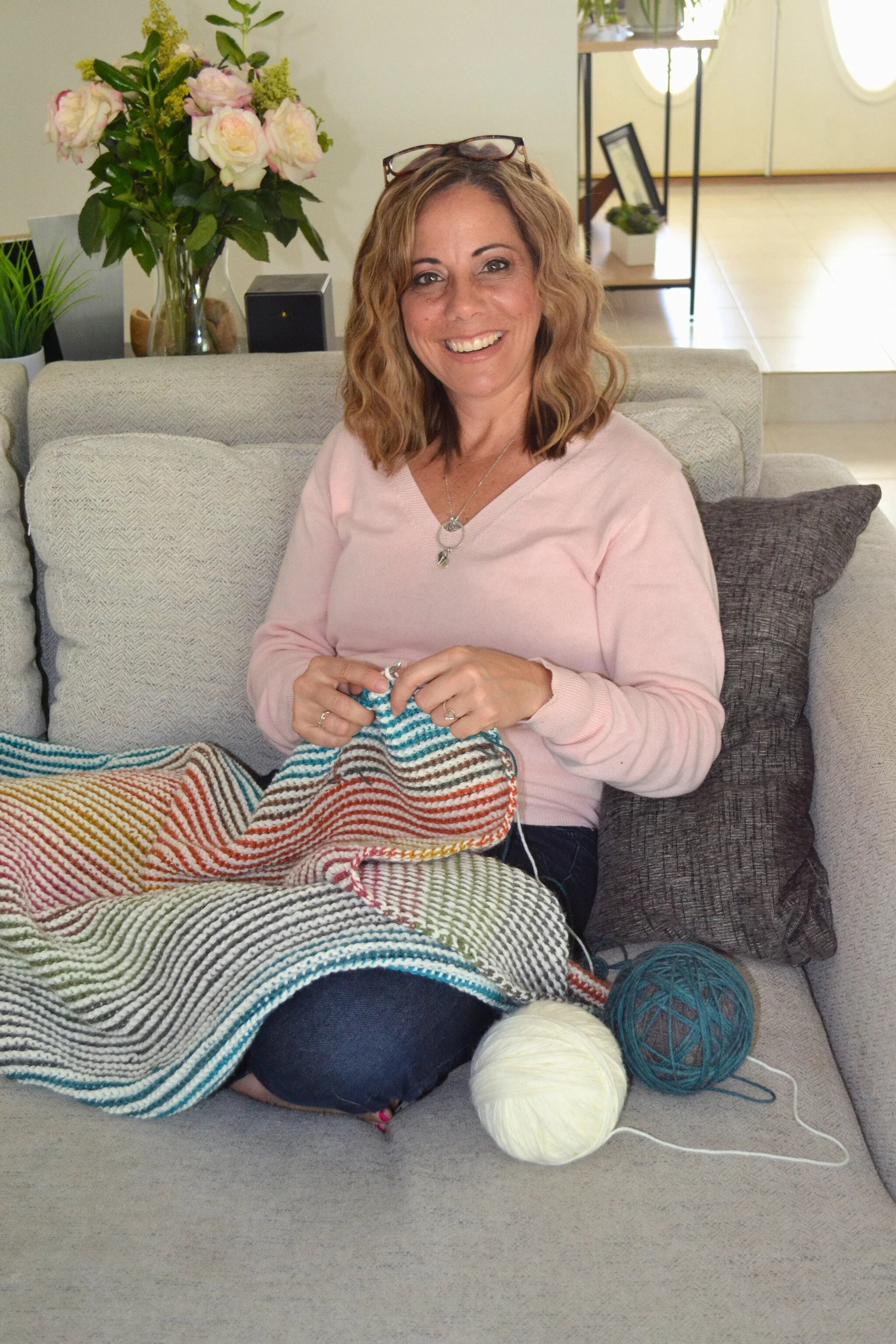 A woman with wavy, shoulder-length hair, wearing glasses on her head, a pink sweater, and a necklace, is sitting on a light gray couch with a gray pillow behind her. She is smiling and knitting a colorful striped blanket with balls of white, cream, and blue yarn on her lap. The background features a vase with pink and white flowers on a table, a black speaker, and houseplants.