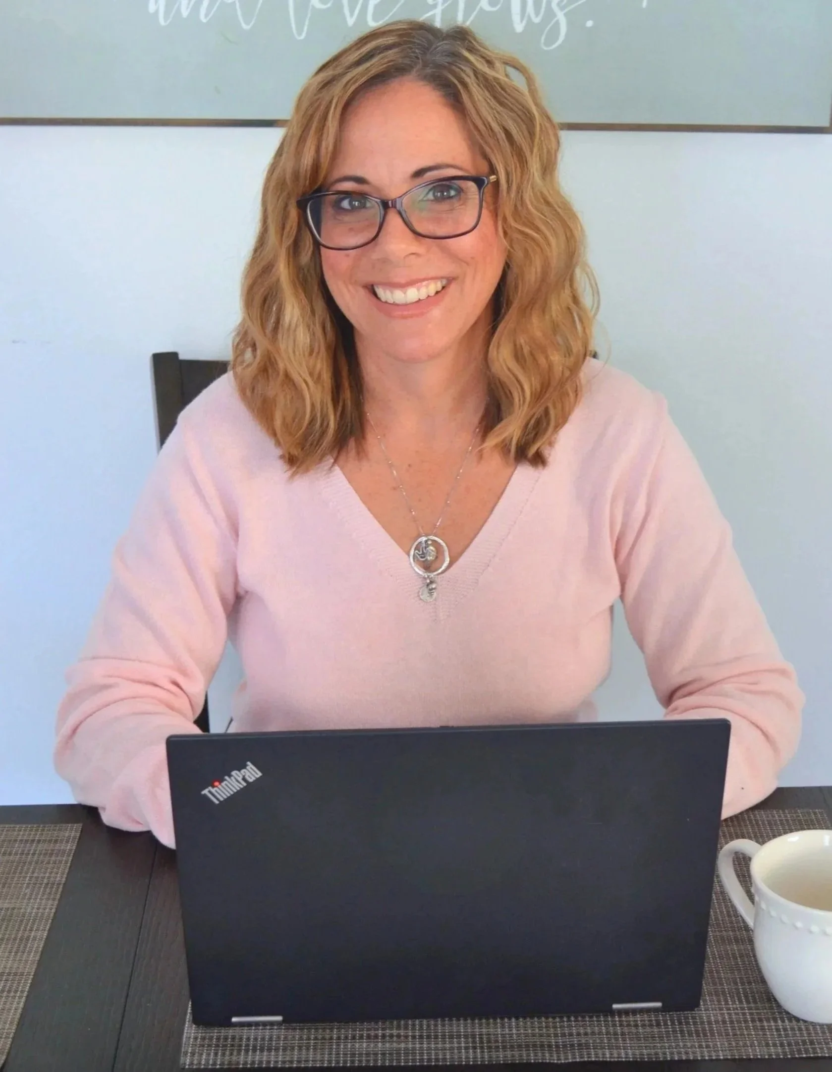 A woman with shoulder-length wavy blonde hair, wearing glasses and a light pink sweater, sitting at a table with a black ThinkPad laptop in front of her and a white mug to her right, smiling at the camera.
