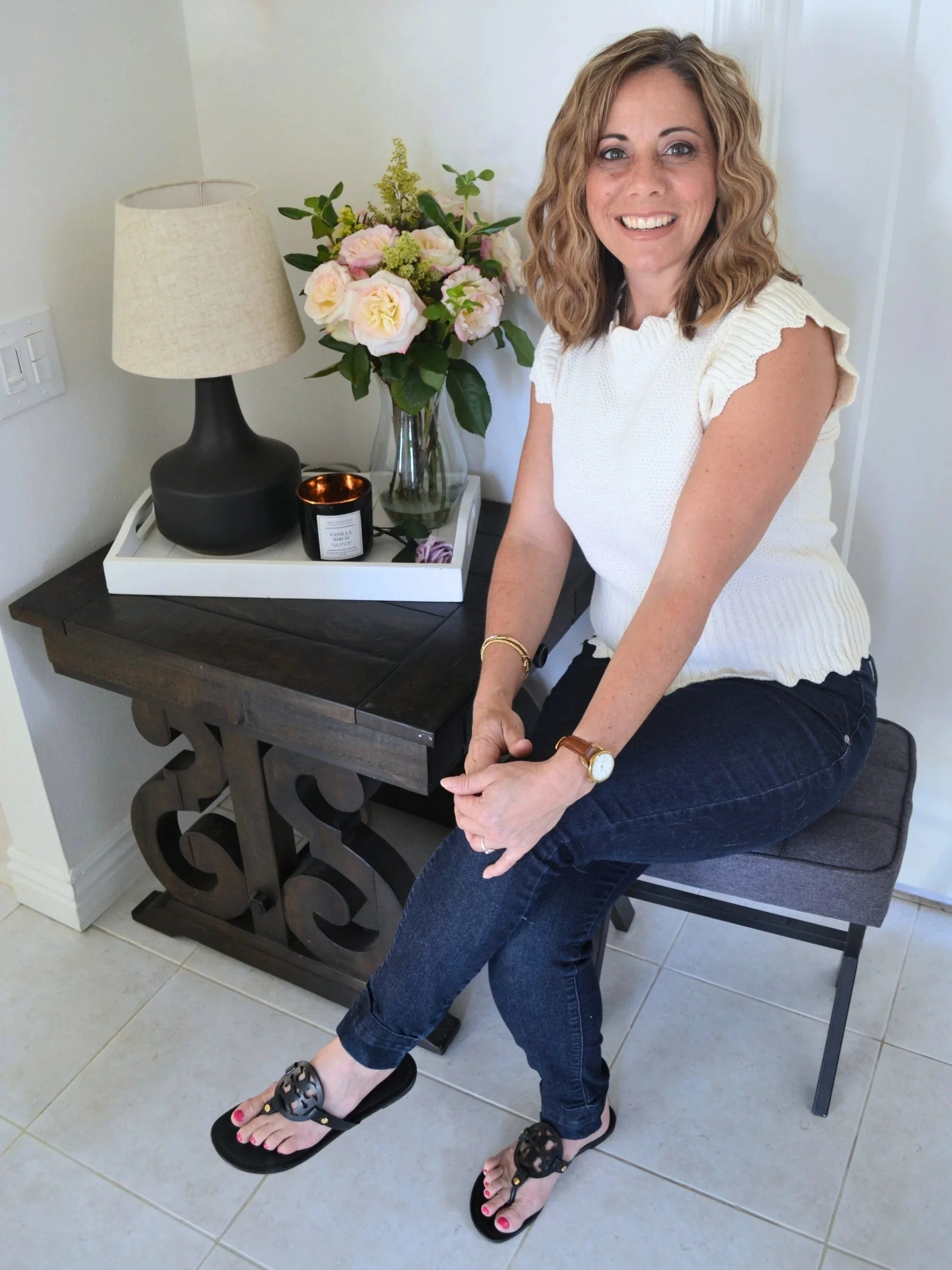 A woman with blonde curly hair and a white knit top is sitting on a chair, smiling. She is wearing dark jeans, a watch, and sandals, and is positioned next to a small black table with a lamp, a vase of pink and cream flowers, a candle, and a small purple item.