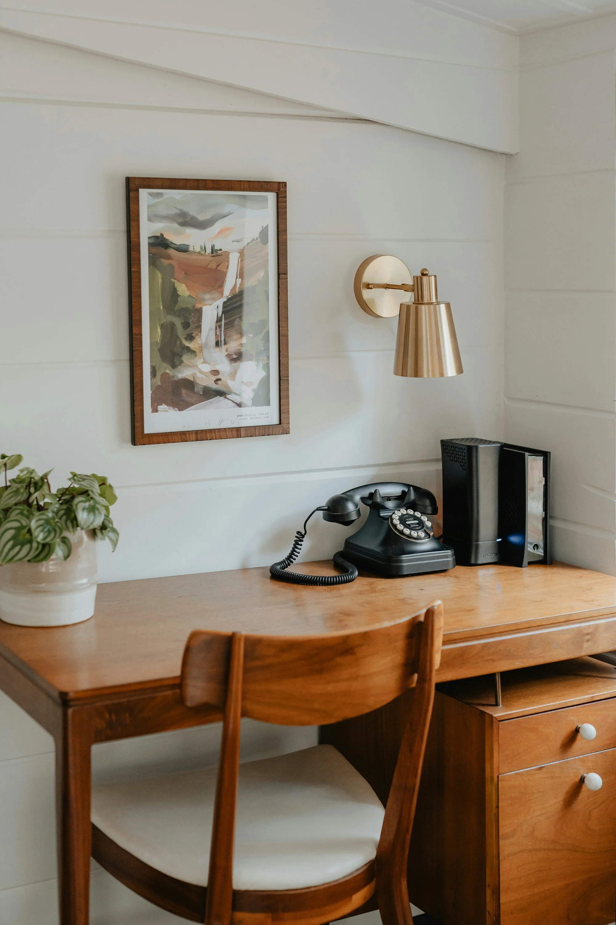 A vintage black rotary telephone, a small stack of black and white binders, and a pot with a green leafy plant on a wooden desk. There is a framed landscape painting on a white wall above the desk, with a brass wall lamp beside it.