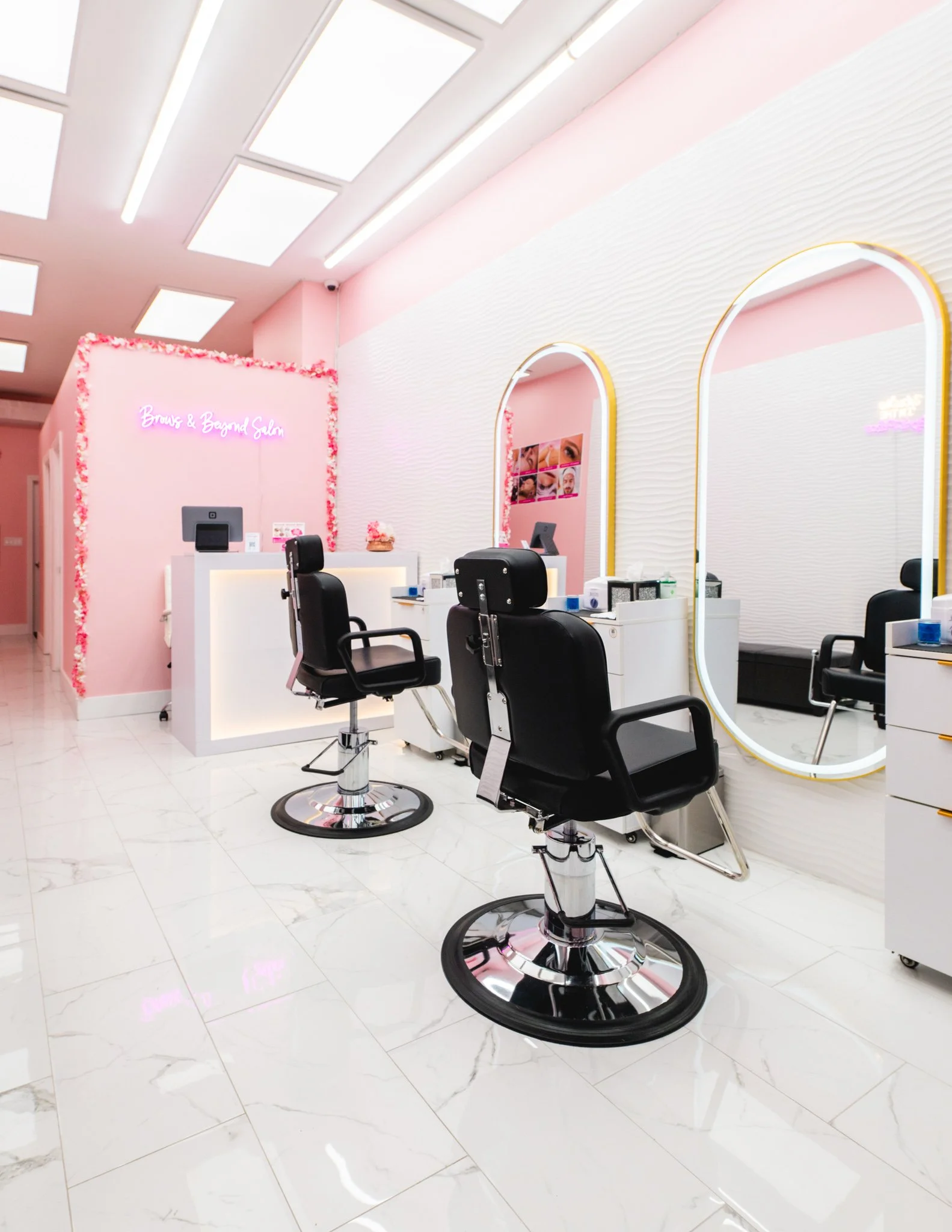 Interior of a nail salon with pastel pink walls, white textured wall accents, oval mirrors outlined in light wood, black salon chairs, and a pink neon sign that reads 'Brows & Beyond Salon' decorated with pink flowers. The salon has white marble flooring and white ceiling panels with lighting.