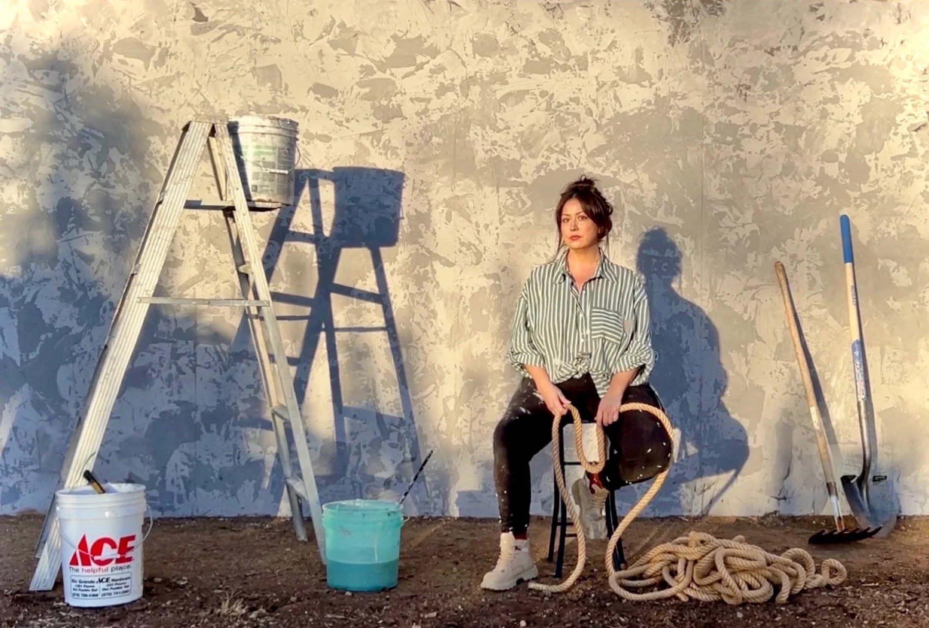 A woman sits on a black chair holding a basketball and a rope, with dry earth floor, against a textured wall. To her left is a stepladder with a paint bucket on top, and two buckets are on the ground. To her right, there are ropes and handles leaning against the wall.