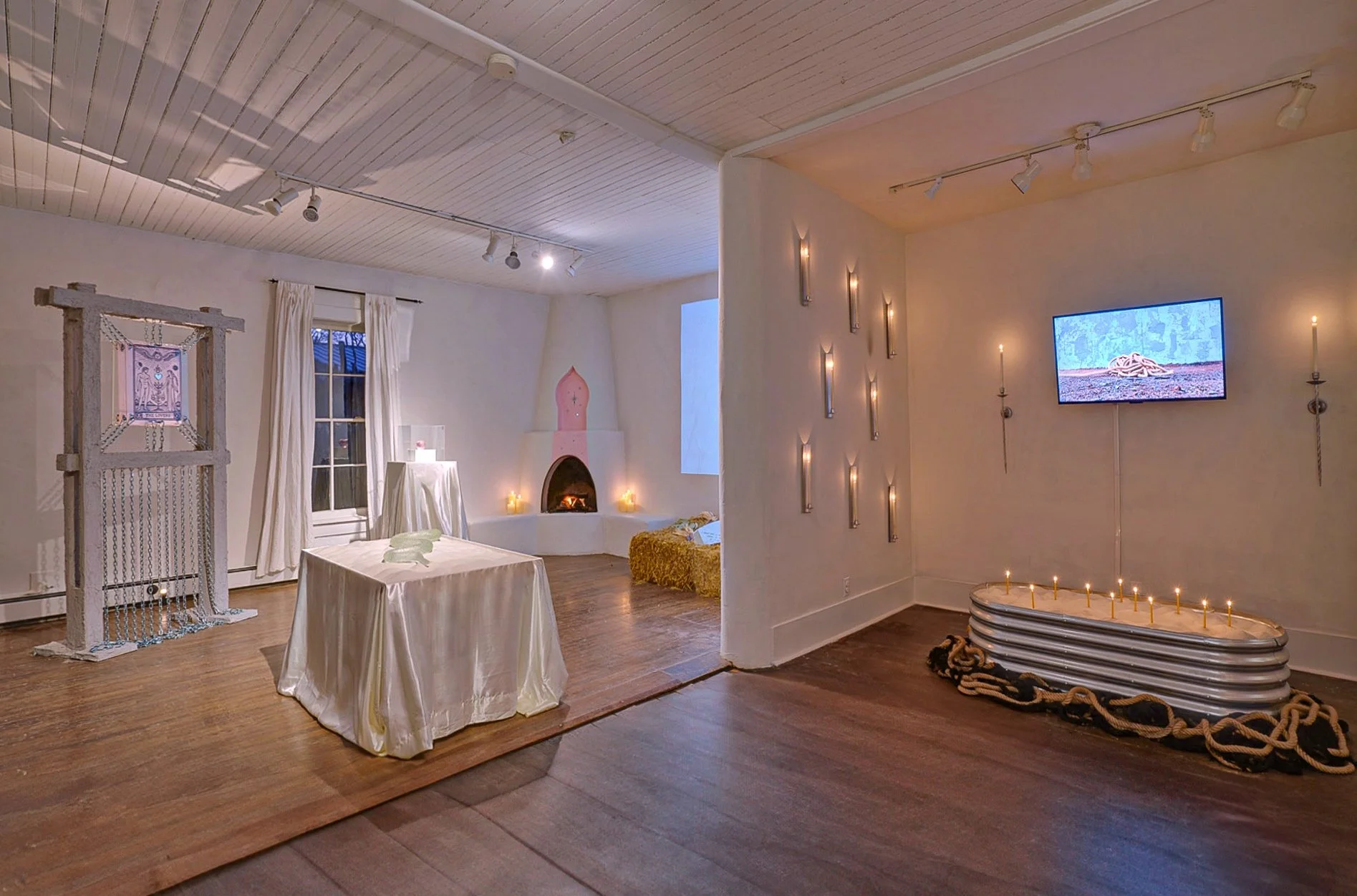 Interior of a room decorated with candles on the walls and a television on the right showing an outdoor scene. There's a fireplace, a window with curtains, and a piece of furniture covered with a cloth. Additional elements include a wooden frame with chains, a table with a cloth, and a rounded object with stacked layers and rope around it.