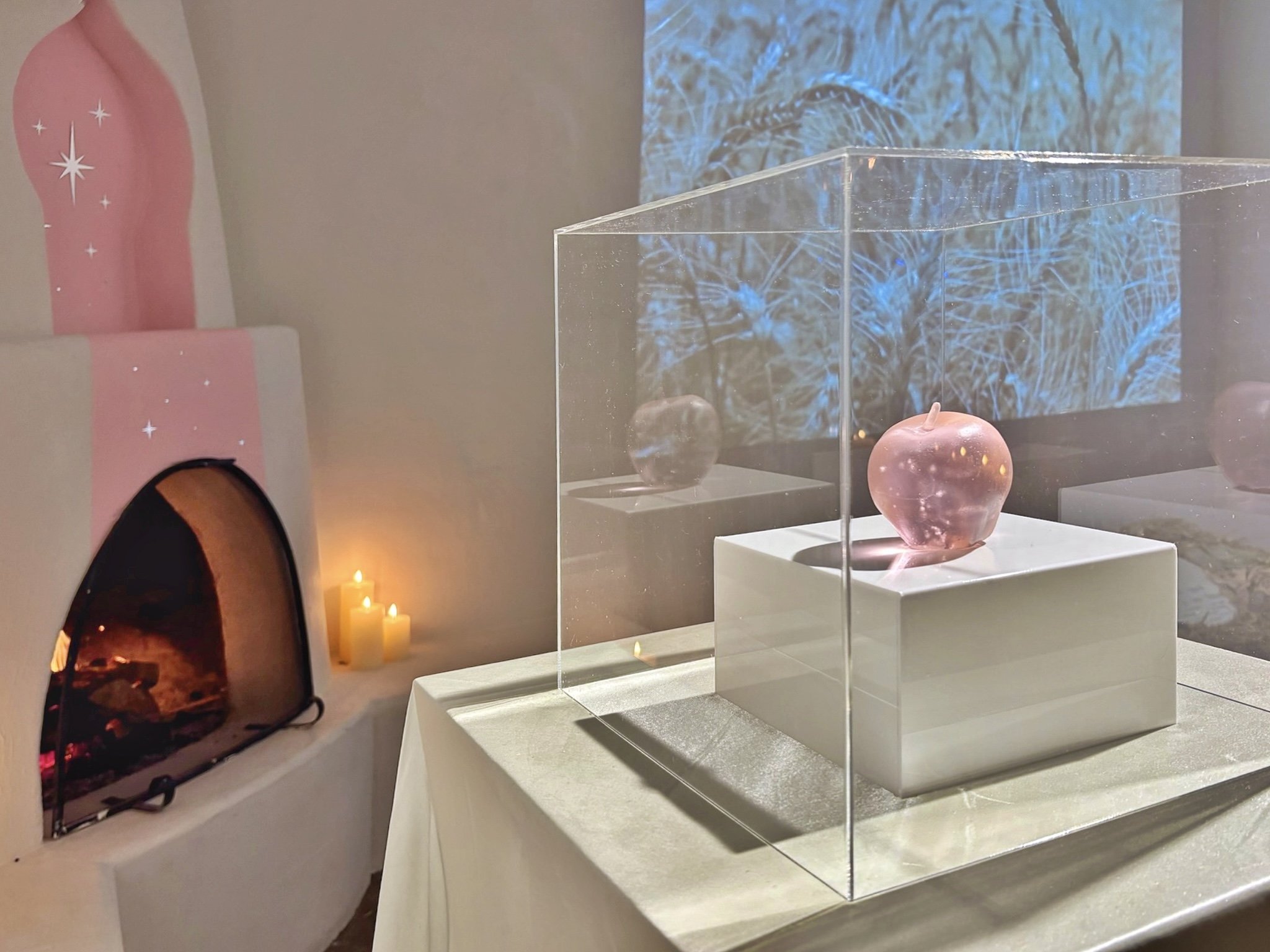 A glass display case containing a pink apple with a small stem in a museum setting. In the background, there is a digital display showing a blue, icy or glass-like texture. To the left, there's a pink decorative item with white stars and a candle beside it.