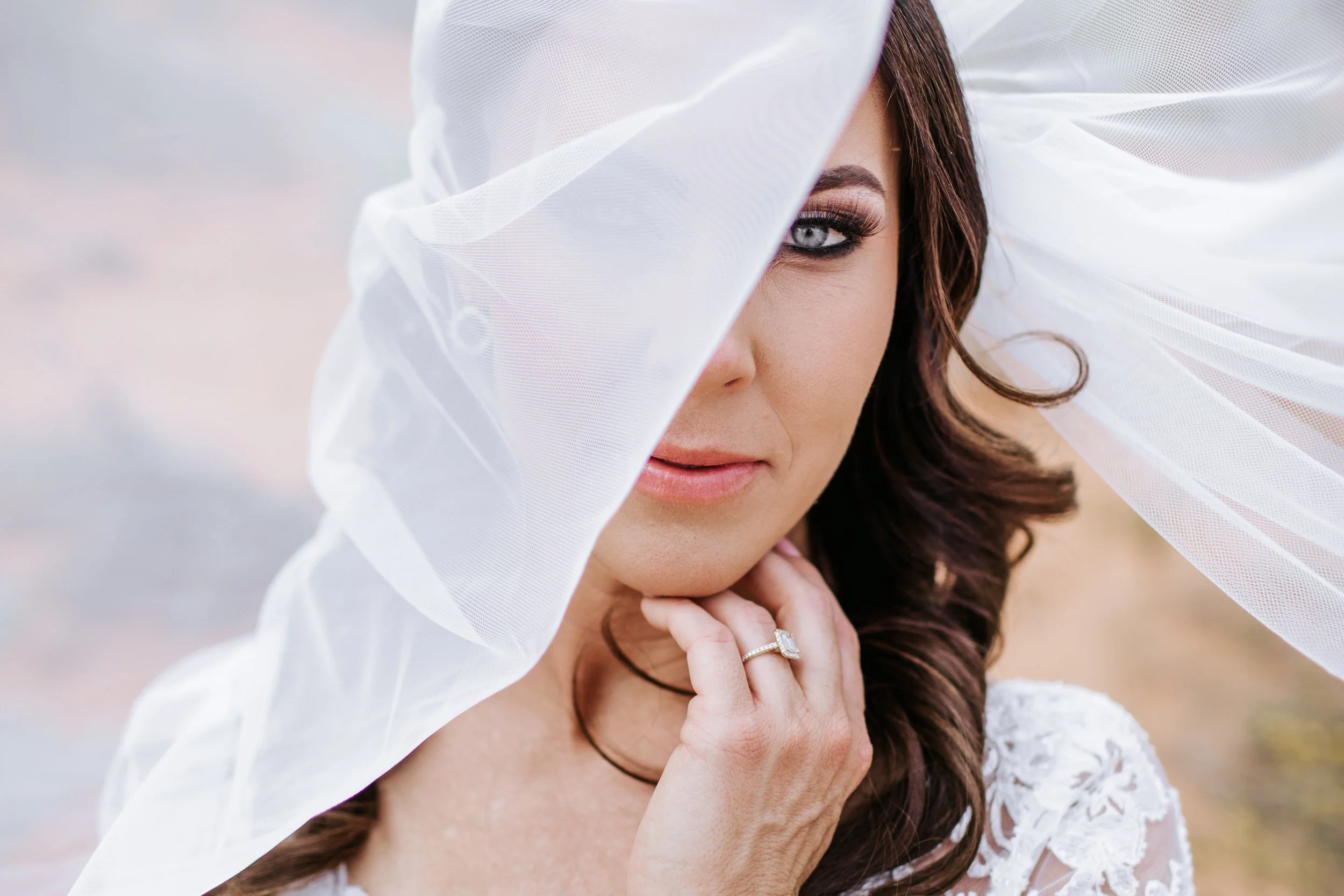 Close-up portrait of a woman with brown hair, blue eyes, and makeup, partially covered by a white tulle veil, wearing a lace dress and a diamond ring, outdoors during daytime.