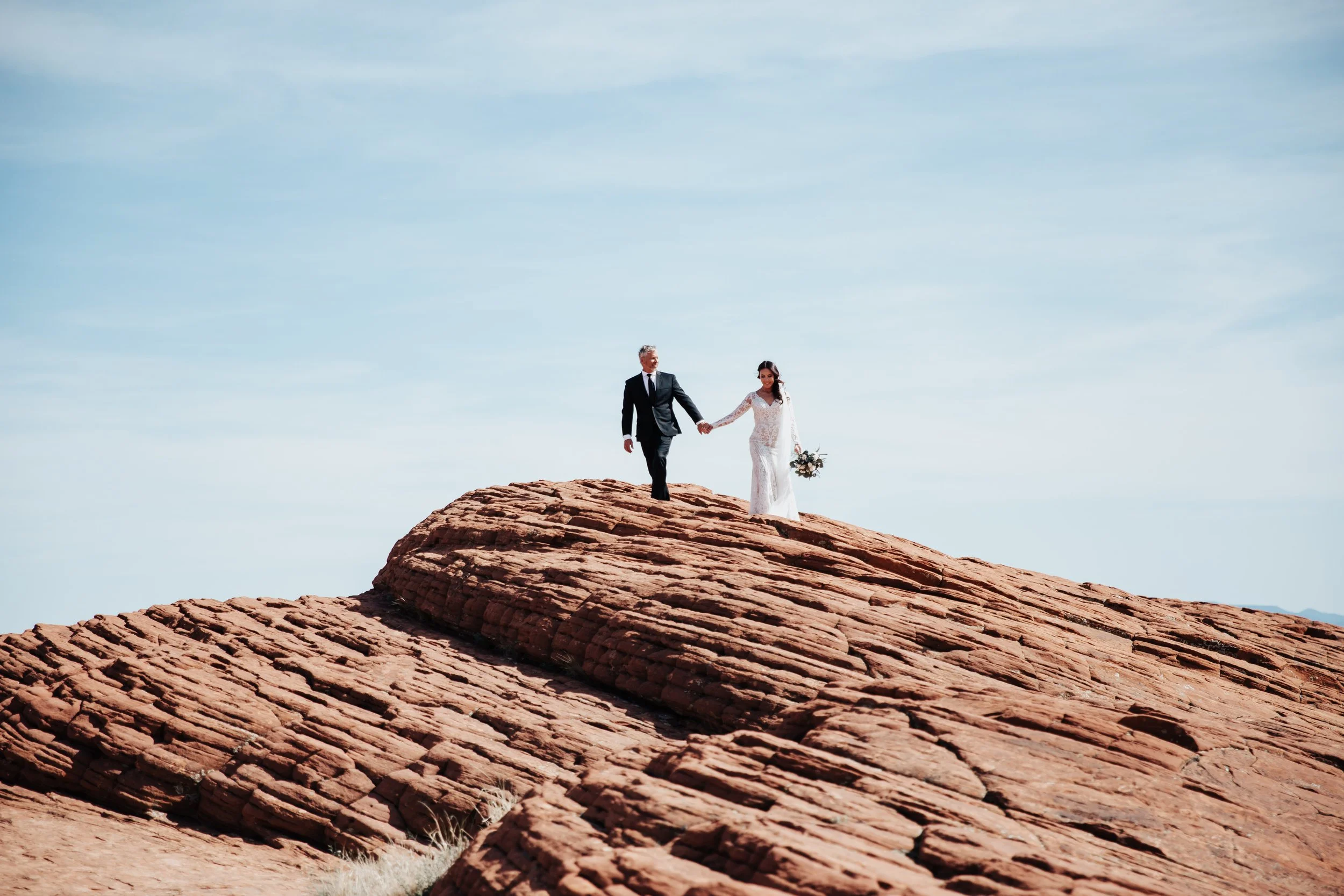 A bride and groom holding hands while walking on large, layered red rocks under a clear blue sky.