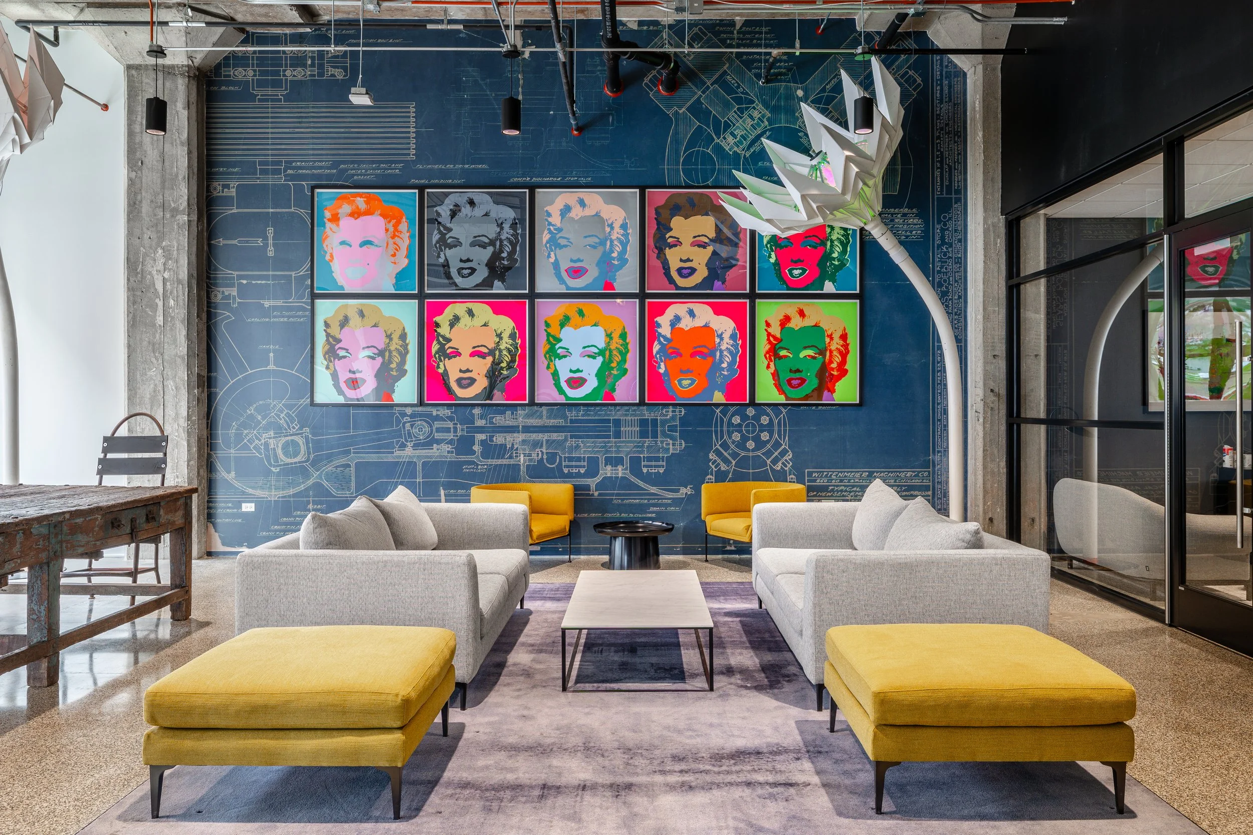 Modern lounge area with light gray sofas, yellow ottomans, and a central white coffee table. The back wall features blueprints and a grid of nine colorful pop art portraits of Marilyn Monroe. A unique white sculptural chandelier hangs overhead, and a