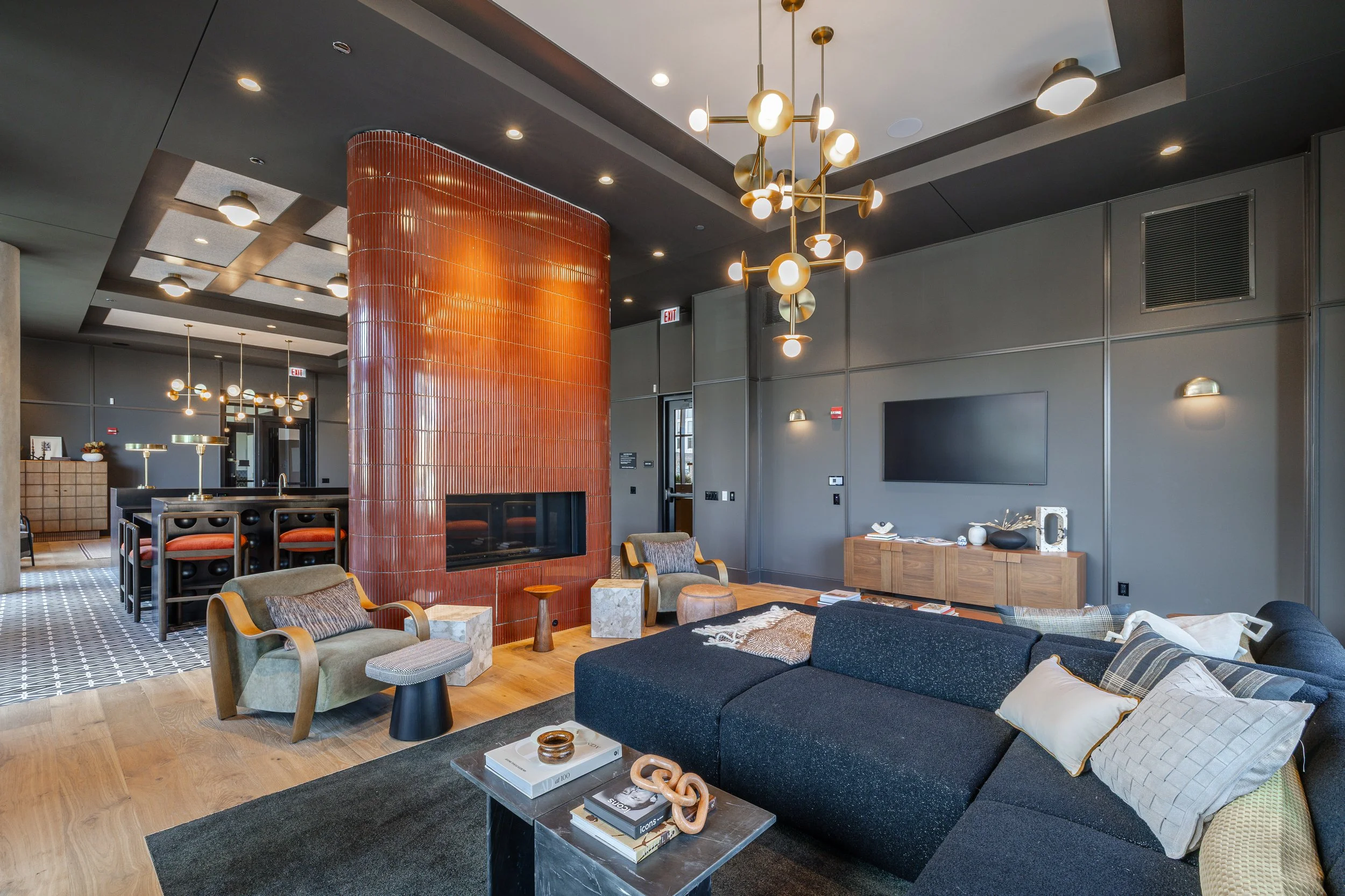 Modern living room with a black sectional sofa, decorative pillows, a coffee table with books and ornaments, a gray armchair with a footrest, a red tiled fireplace, and a minimalist wooden media console, all illuminated by contemporary pendant and wa