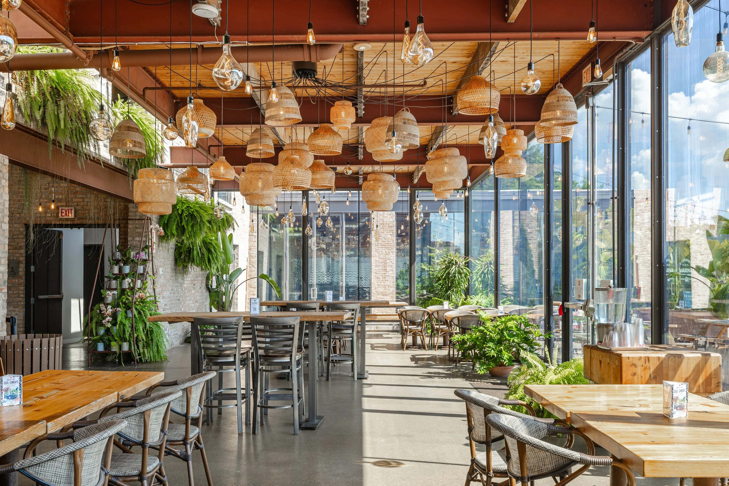 Interior of a bright, modern café with large glass windows, hanging wicker and glass light fixtures, wooden tables, and green plants.