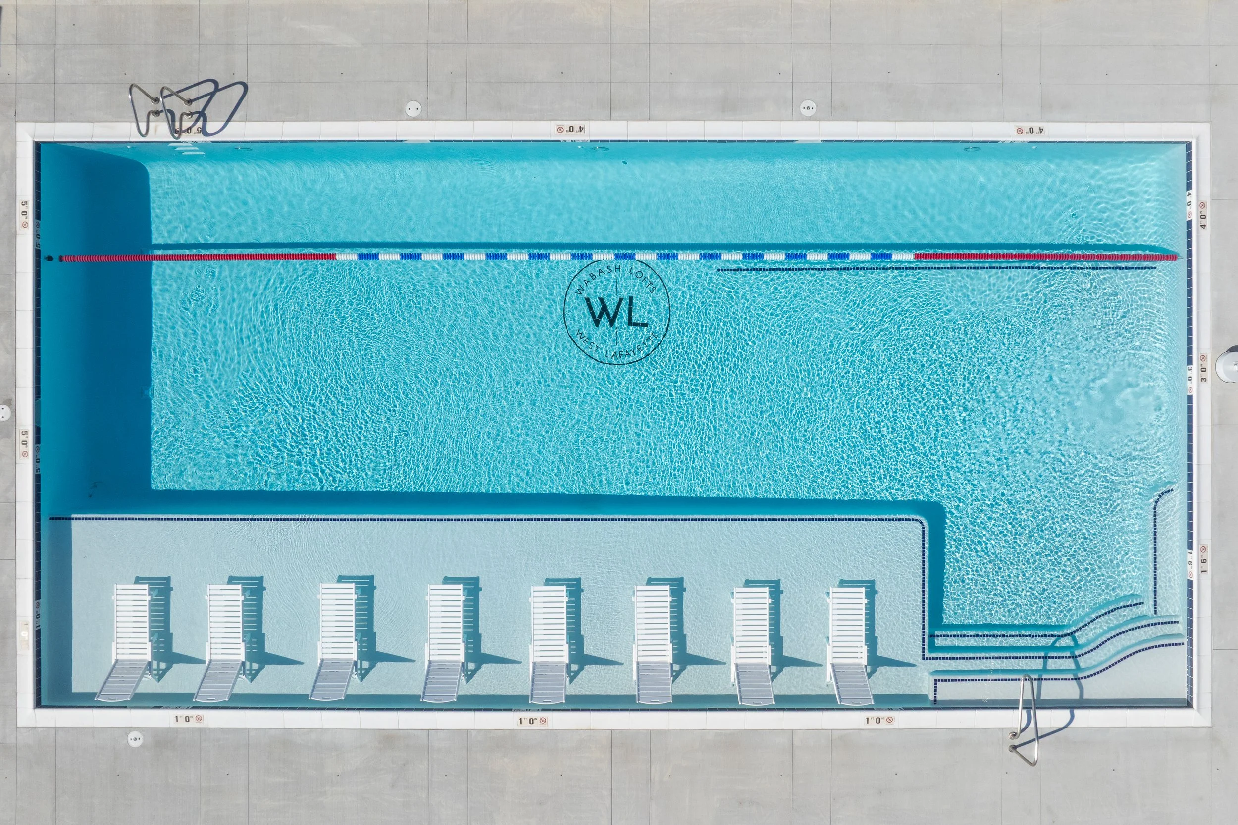A rectangular outdoor swimming pool with a set of six white lounge chairs along one side. The pool has blue water and is surrounded by a concrete deck. It features a black and white lane divider, stairs on one end, and a logo at the center that reads