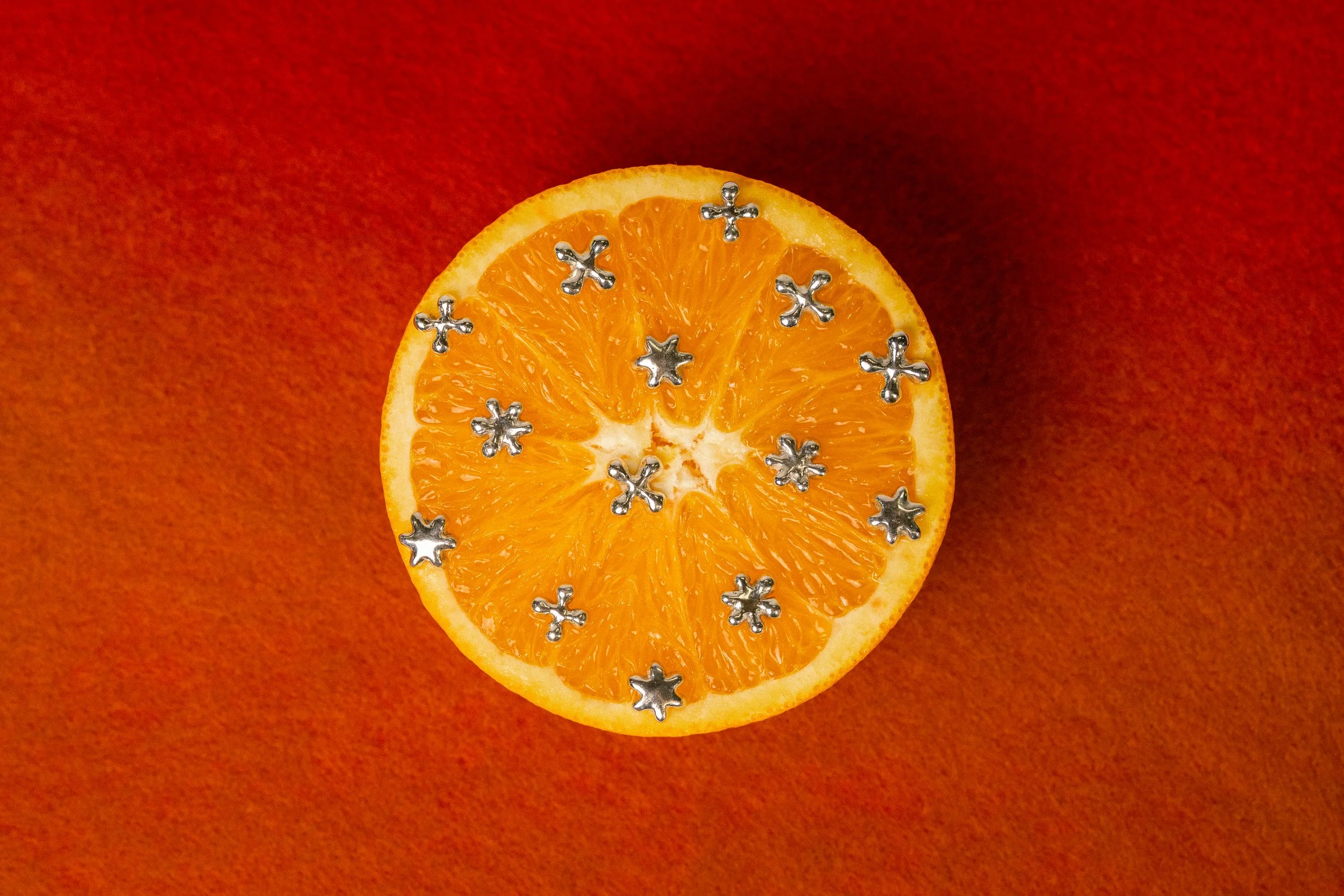 A halved orange with small metallic star-shaped and flower-shaped charms on its surface, placed on an orange background.