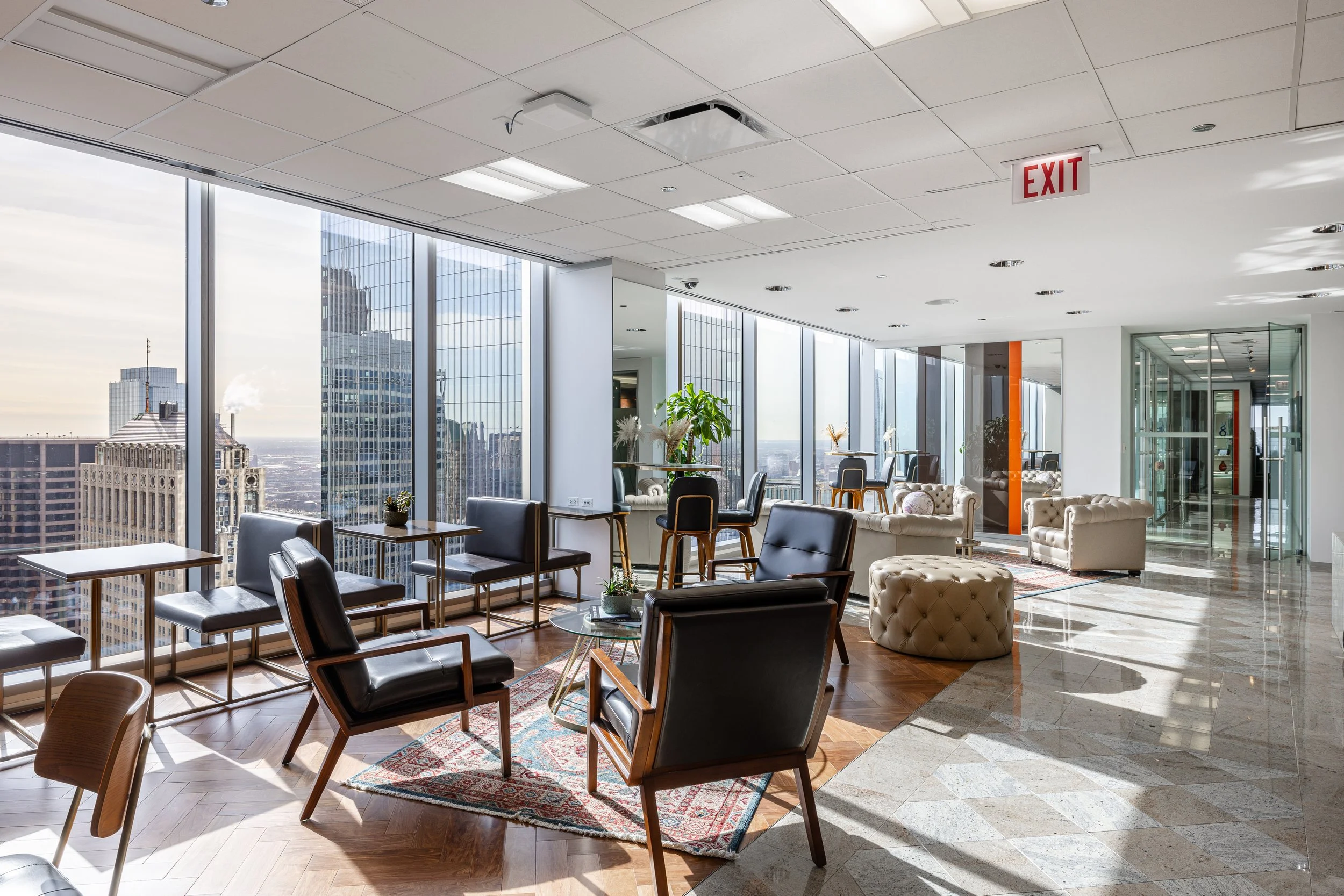 Modern office lounge with mid-century style chairs, sofas, glass tables, large windows with city skyline view, potted plants, and natural sunlight.