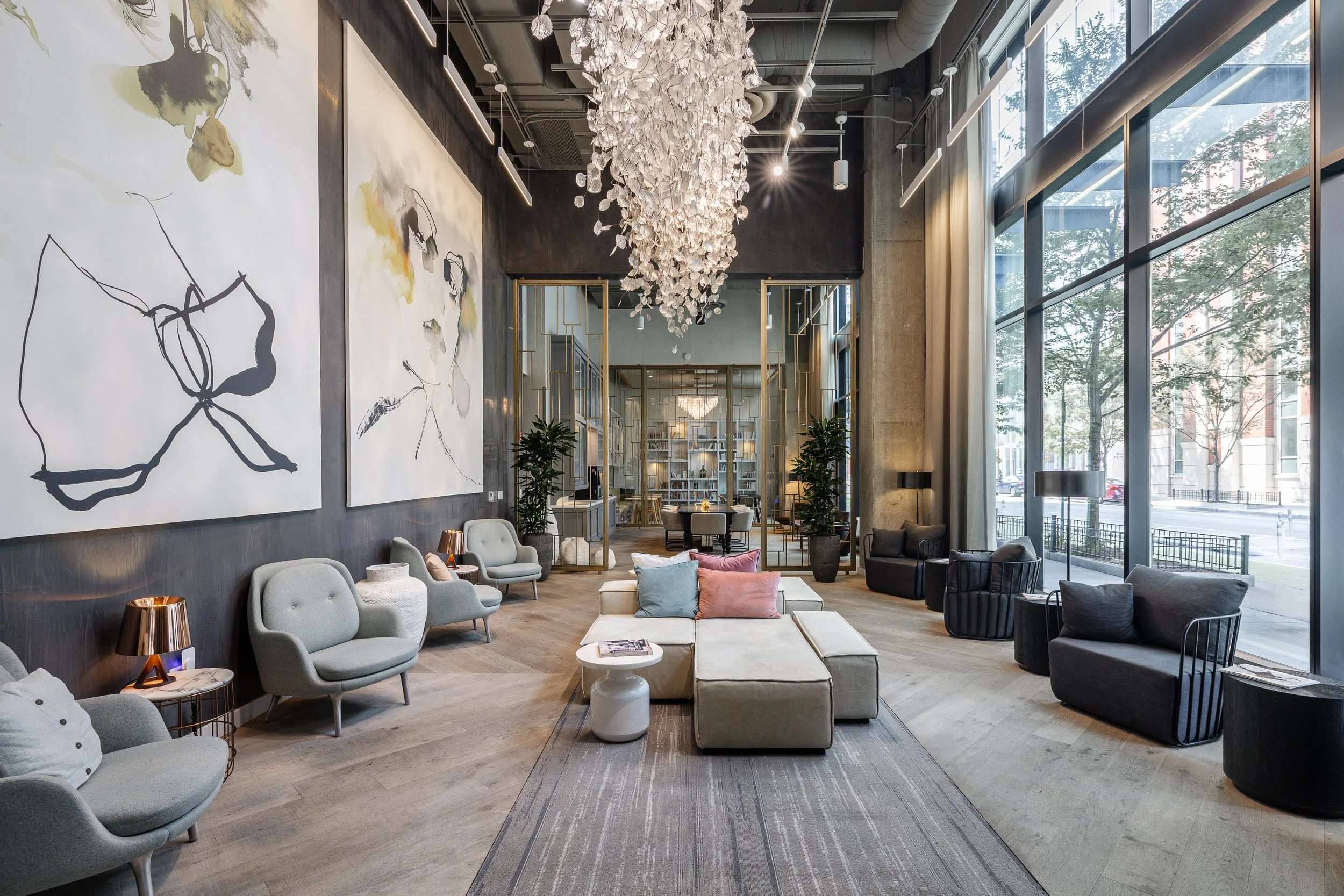 Modern hotel lobby with large floor-to-ceiling windows, stylish seating, contemporary art on walls, a white chandelier, and a library area in the background.