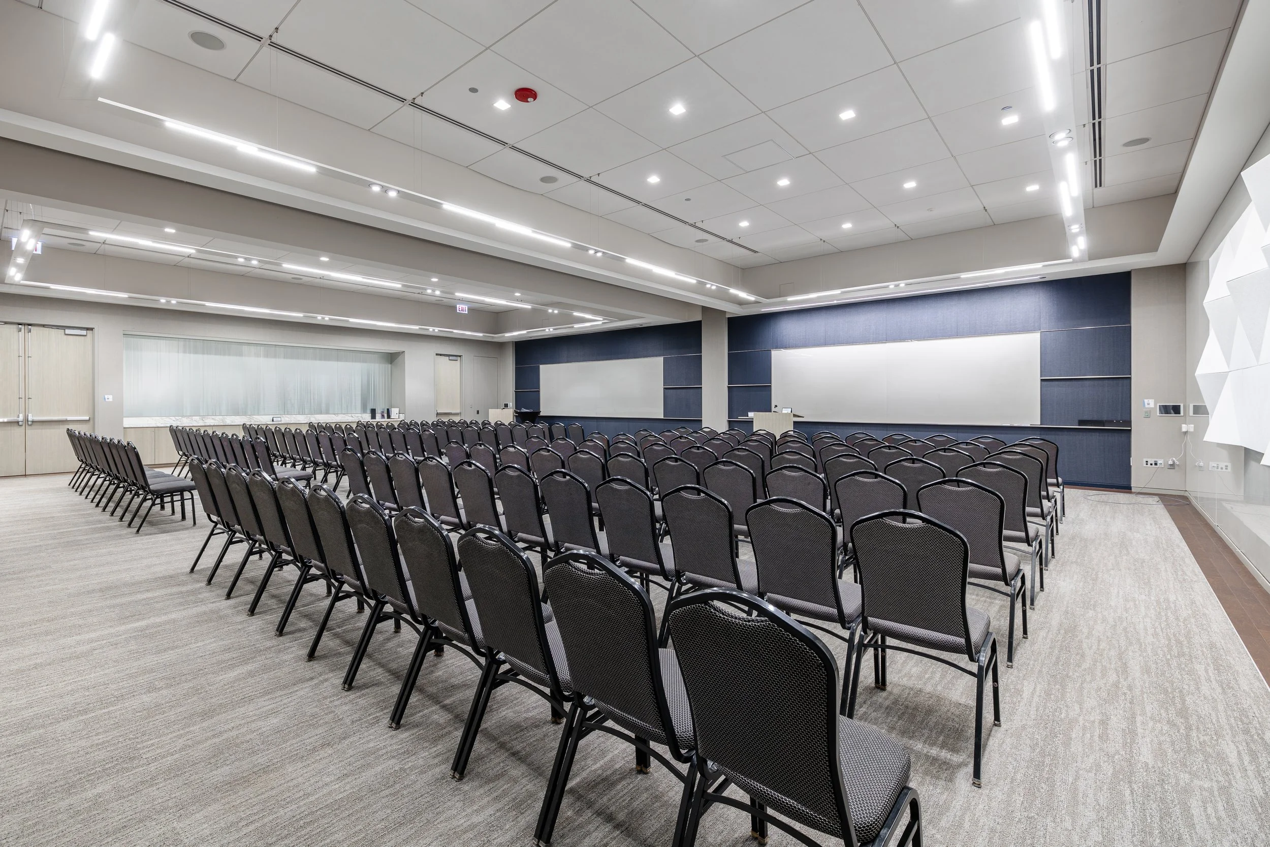Empty conference or presentation room with rows of black chairs facing a whiteboard and podium, ceiling with bright lighting.