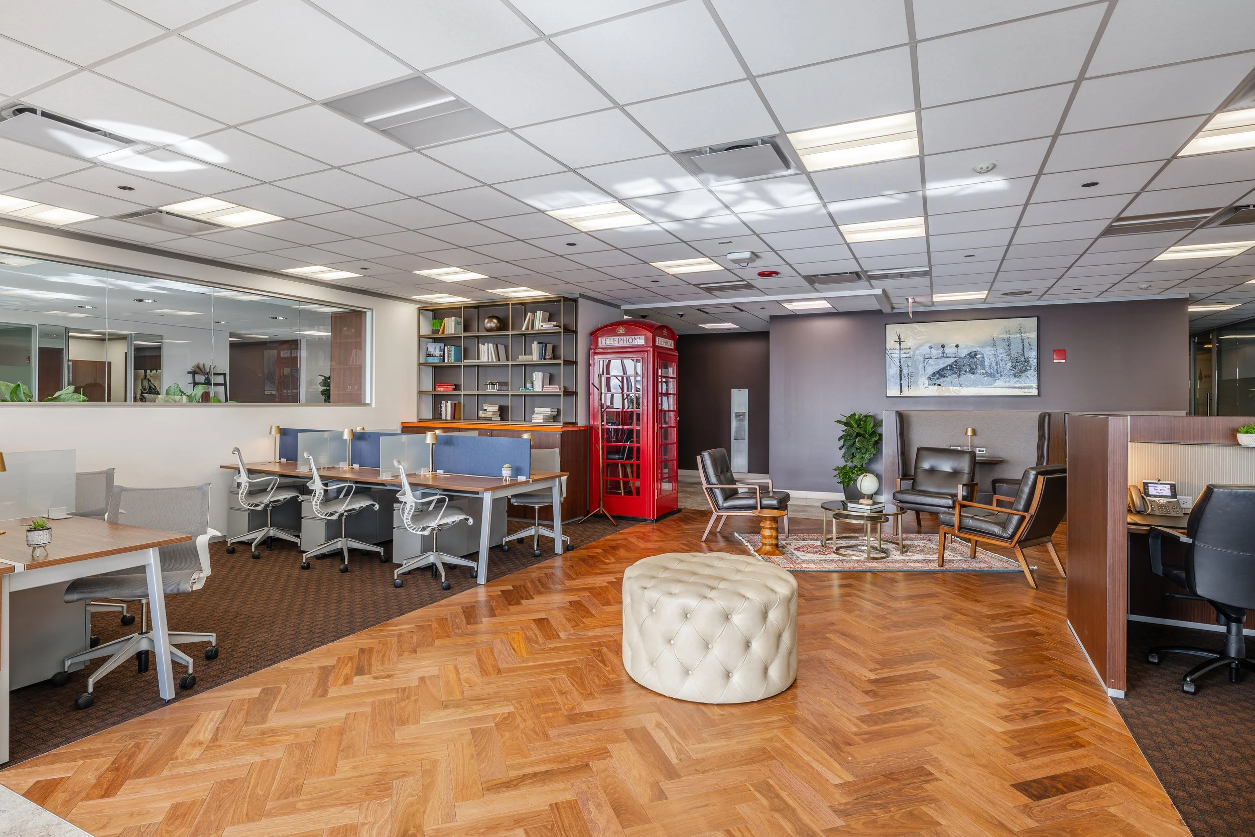 Modern office lounge with wooden flooring, leather chairs, a vintage red telephone booth, a bookshelf, and a large windowed wall.