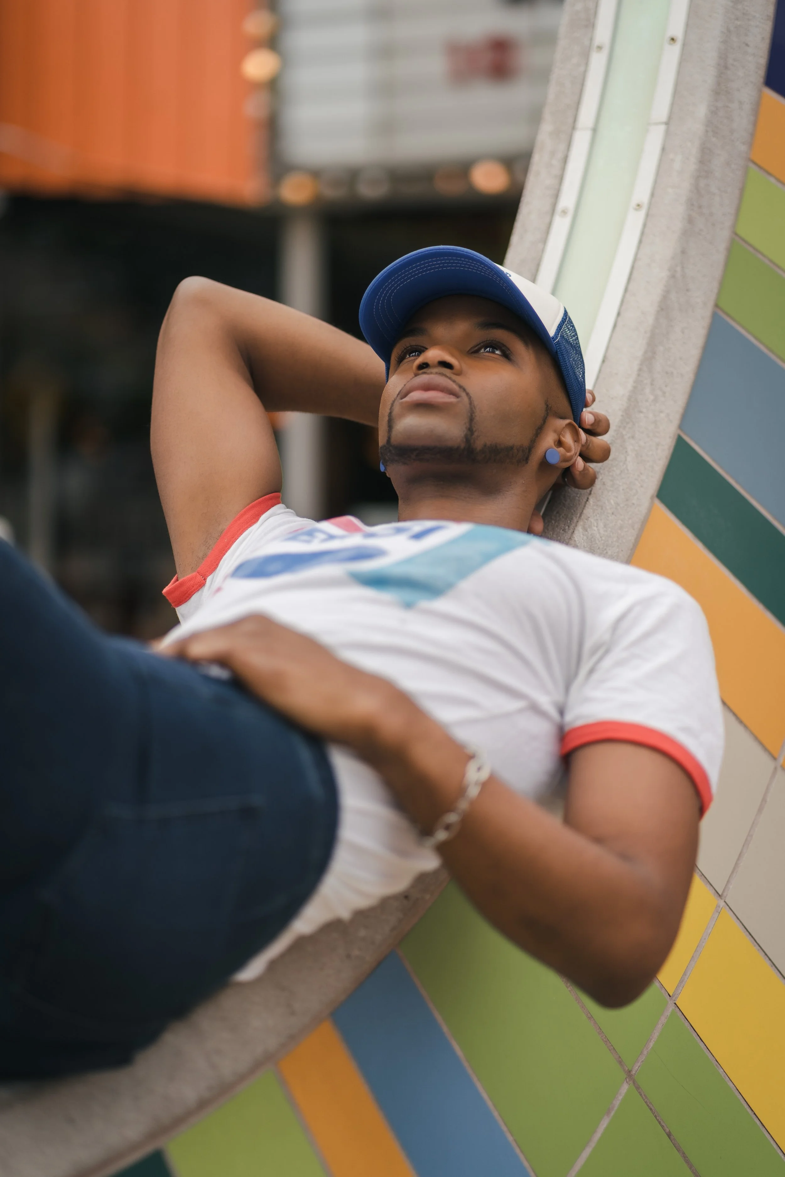 Young man relaxing with his hands behind his head, lying on a colorful tiled ledge, wearing a baseball cap and casual clothes.